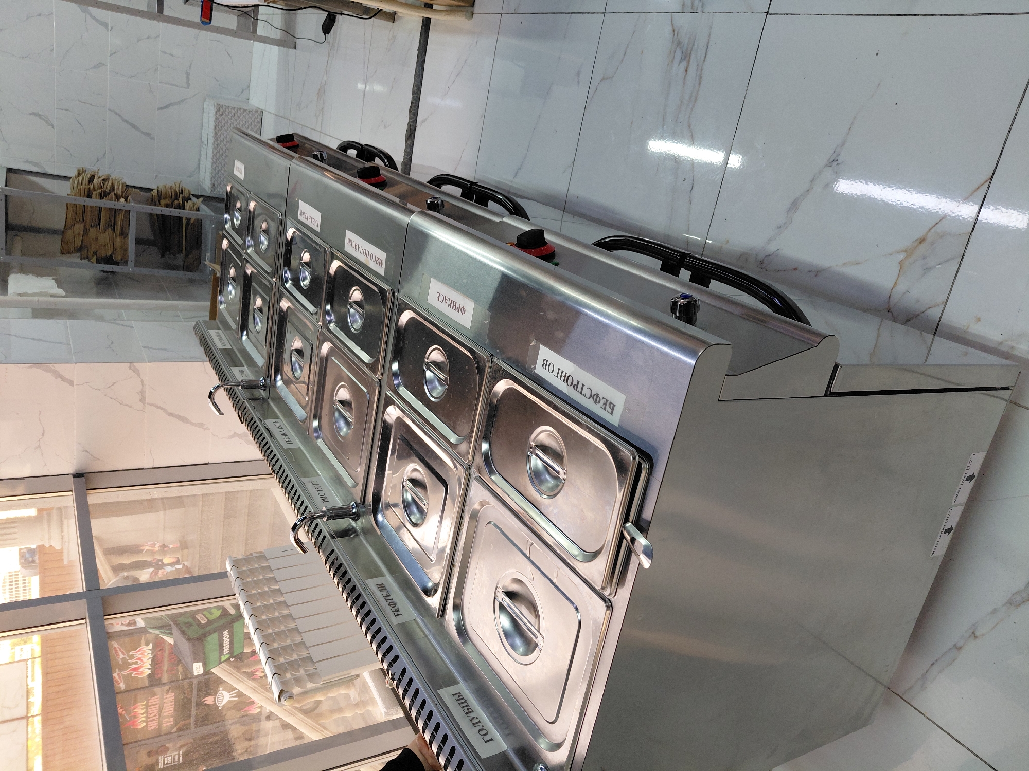 This is a tilted, eye-level, outdoor shot of a bain-marie food warmer unit in what appears to be a restaurant or food service establishment in Aktau, Kazakhstan. The unit is made of stainless steel and has multiple compartments with lids, each labeled with Russian text, likely indicating the food items they hold. Visible labels include "ТЕФТЕЛИ" (Meatballs), "ГОЛУБЦЫ" (Cabbage Rolls), "БЕФСТРОНГОВ" (Beef Stroganoff), "ФРИКАСЕ" (Fricassee), and "МЯСО ПО-ТАЙСКИ" (Thai Meat). The unit has black handles and red control knobs. In the background, through a glass window, reflections of people and various food-related graphics are visible, including what looks like shashlik and a graphic with flames. To the left of the bain-marie, there are racks holding what appear to be bundles of dried goods, possibly herbs or spices. The floor is tiled with a marble-like pattern. The lighting suggests daytime.