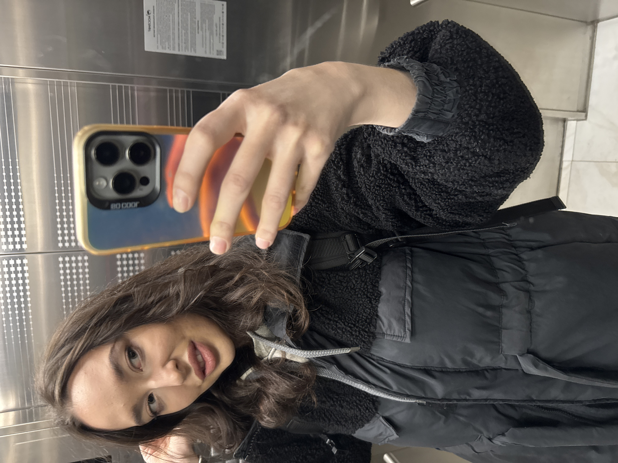 A young woman with long, wavy dark brown hair and light-colored eyes is featured in a selfie. She has full lips with a subtle pink hue and a calm, slightly smiling or pouting expression. She is dressed in a black jacket, which appears to be a puffer style combined with fuzzy, sherpa-like material on the shoulders and arms, and has gathered elasticated cuffs. A black strap, possibly from a bag or part of her jacket, crosses her chest.
She is holding a smartphone, which is angled towards herself for the selfie. The phone, identifiable as an iPhone due to its triple-lens camera module, is encased in a light-colored (beige/golden) case featuring a graphic of a planet or celestial body with gradients of orange and blue. The text "BO COOF" is visible on the phone case.
The setting appears to be an indoor environment, likely an elevator or a modern hallway, characterized by metallic, reflective wall panels. These panels have distinct vertical grooving and patterns of small square indentations. On an upper metal panel in the background, a white rectangular sticker or sign is visible. Prominently displayed on this sign is the word "ТИНЬКОFF" (TINKOFF), a Russian bank, along with smaller, illegible text. The image does not provide clues about the time of day or weather.