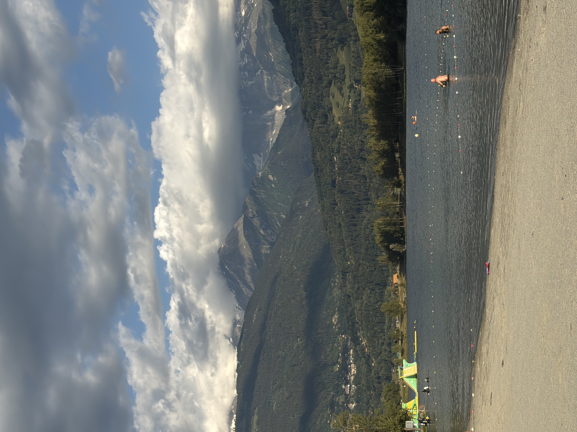 The image depicts a summer day at the Pas de Cheville lake in Passy, France. In the foreground, a sandy beach is visible with some scattered debris. The lake occupies the midground, with several people swimming or wading in the water. A bright yellow inflatable water slide structure is present near the shore on the left side of the image, with some people near it. Buoys mark off swimming areas in the lake. In the background, a densely forested mountain slopes upward, topped by snow-capped peaks partially obscured by dramatic, cloudy skies. The overall scene conveys a sense of leisure and outdoor recreation against a majestic natural backdrop.