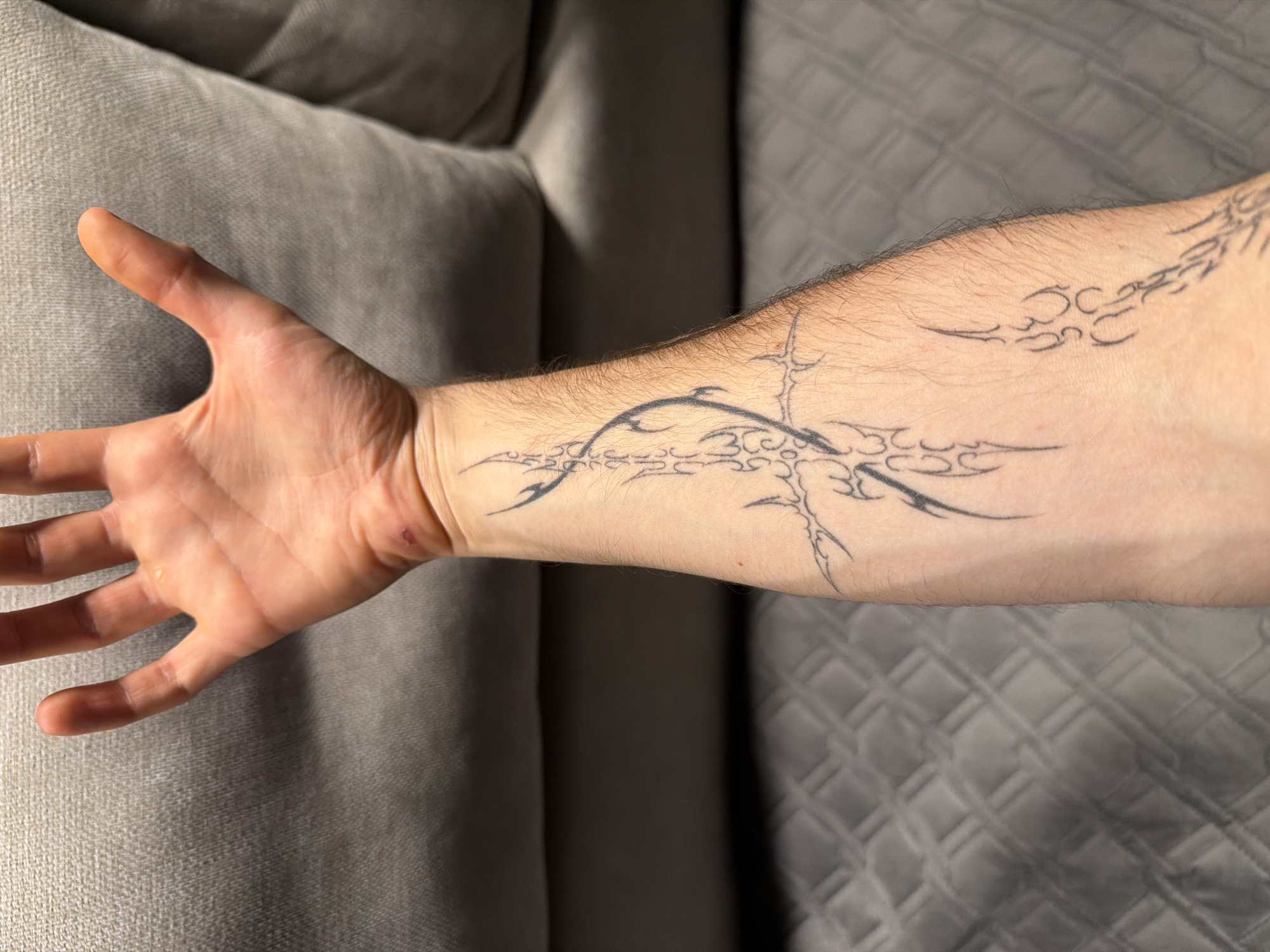 An overhead shot shows a person's left arm with a black tribal-style tattoo. The arm is resting on a grey quilted surface, possibly a couch or blanket. The tattoo begins near the wrist and extends up the forearm, featuring intricate, flowing lines and sharp points. The person's left hand is visible at the top of the frame, palm facing upwards, with fingers slightly spread. The background consists of a grey textured surface, likely a sofa cushion. The scene is indoors and appears to be a casual setting. The image does not provide any information about the city of Almaty, Kazakhstan.