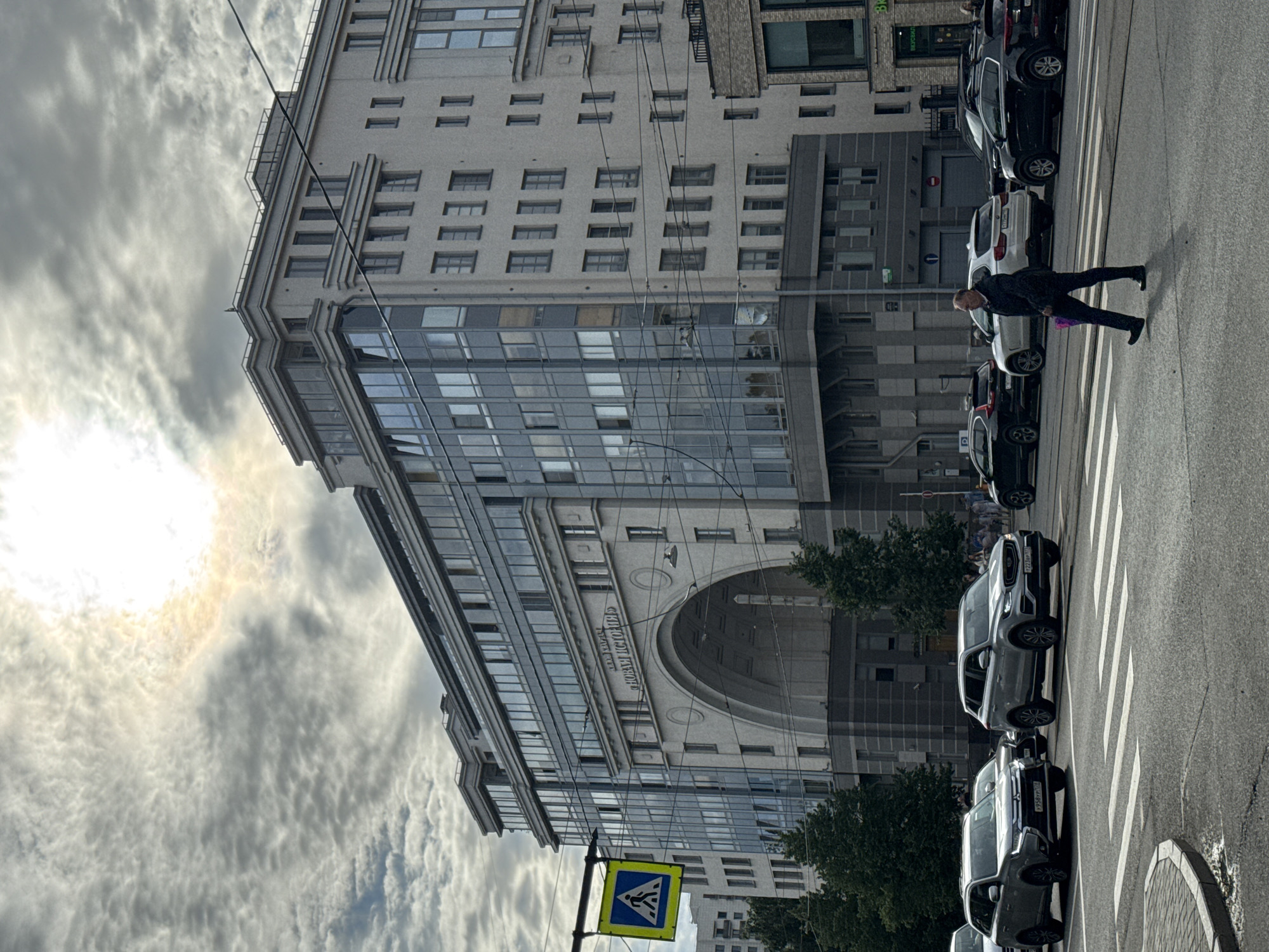 A man in a dark suit and dark pants is walking across a pedestrian crossing on a street in Saint Petersburg, Russia. He is carrying a small, purple bag in his right hand. The man is crossing in front of several cars, including a silver SUV, a black SUV, and a white SUV. The buildings in the background are modern and tall, with glass windows and white facades. A large arched entrance is visible on the central building, with Russian text above it. The sky is cloudy with the sun peeking through. A yellow pedestrian crossing sign is visible on the left side of the image.
