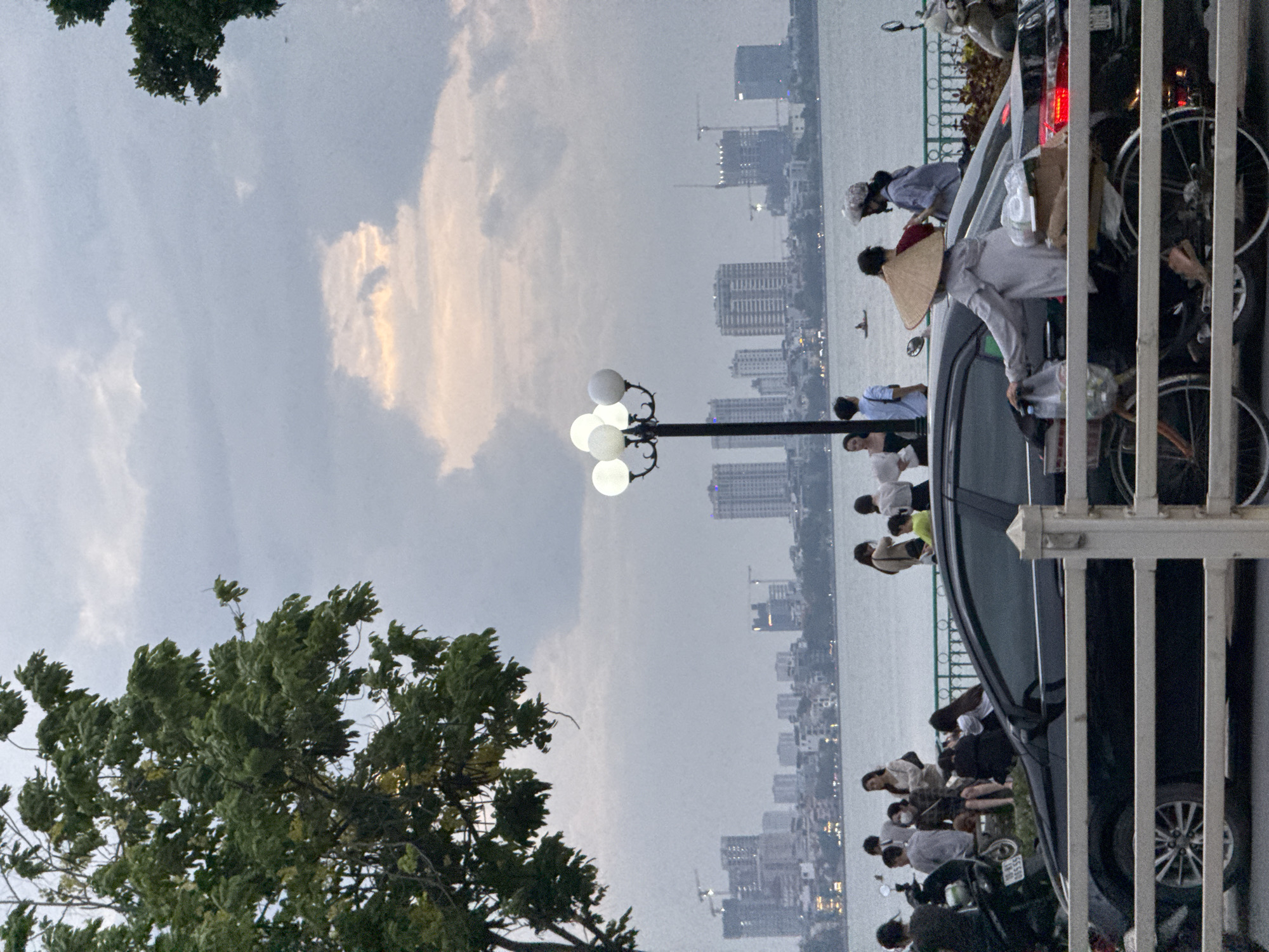 The image depicts a scene by West Lake (Hồ Tây) in Hanoi, Vietnam, under a cloudy sky with hints of sunset colors breaking through. In the foreground, a black sedan is parked, partially obscured by a white railing. To the right of the car, a person wearing a conical hat is pushing a bicycle laden with bags and boxes. Further back, a group of people are gathered near a low green fence overlooking the lake. Beyond the fence and the water, the Hanoi skyline is visible, featuring numerous modern buildings and construction cranes. A street lamp with multiple globes stands to the left of the center, illuminating the dusk. Trees with dense green foliage frame the left side of the image, adding to the natural elements of the scene. The overall atmosphere is that of a relaxed evening gathering by the water, with urban elements forming the backdrop.