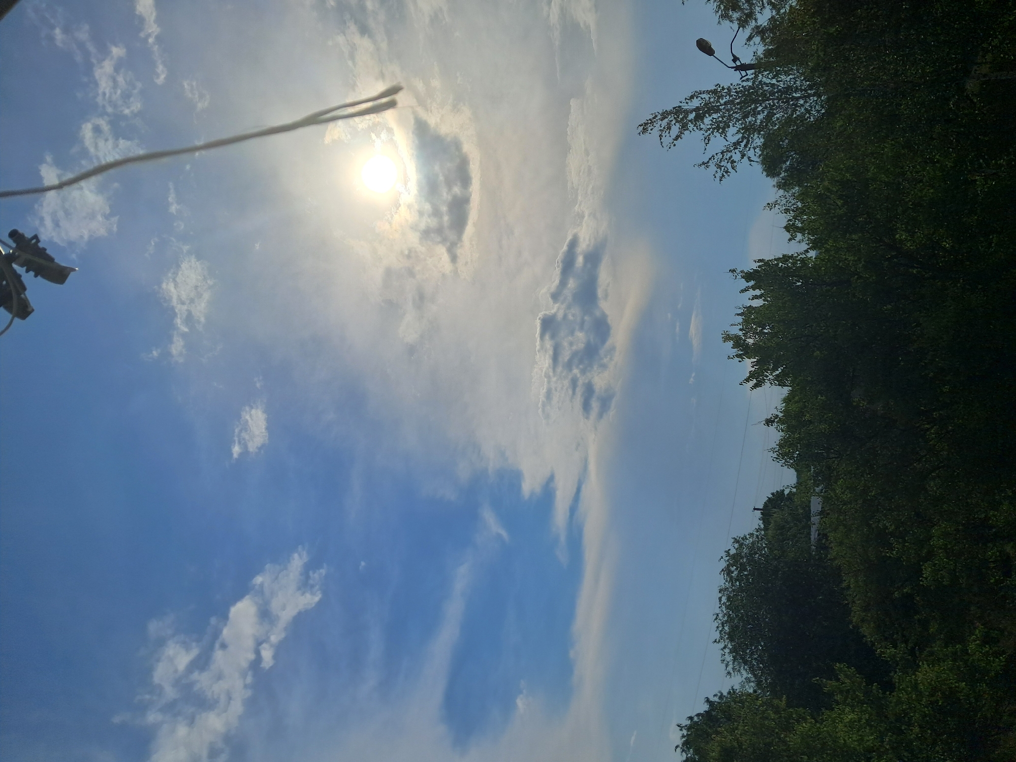 The image captures a view of the sky above a treeline in the city of Smila, Ukraine. The sky is a bright blue with scattered white clouds, and the sun is visible in the upper right quadrant, partially obscured by clouds, creating a bright glare. A thick electrical wire or cable hangs diagonally from the top center of the frame, with what appears to be electrical hardware attached to it. The lower portion of the image is dominated by the dense, dark green foliage of trees, with hints of rooftops and a streetlamp visible through the canopy. The overall impression is a daytime scene, likely during fair weather, with a mix of natural and man-made elements.