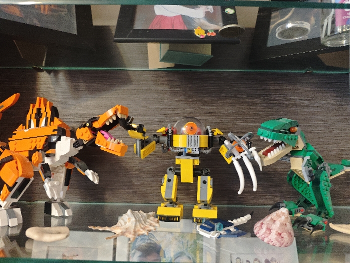 The scene, located indoors in Samara, Russia, depicts a display on a clear glass shelf. Three primary Lego figures are arranged centrally. On the left is a large orange, black, and white striped creature, resembling a sabertooth tiger, positioned with its mouth open, revealing a pink tongue and white teeth. In the center stands a yellow and gray bipedal robot featuring a transparent orange dome head with an internal orange eye. Its left arm, equipped with a yellow claw, is extended towards the tiger-like creature's open mouth. On the right is a green Lego dinosaur, resembling a Tyrannosaurus Rex, also depicted with an open mouth exposing a red tongue and white teeth. Its left arm, terminating in three large white claws, is directed towards the robot.
On the glass shelf below the figures, from left to right, are a small, light-colored ring-shaped object, a large light brown spiral seashell, a miniature blue and white Lego spaceship, a reddish-pink textured rock or coral piece, and a small off-white organic object. The background above the shelf is dark, likely wood, and partially reveals a framed picture containing a cartoonish female figure, along with other miscellaneous objects and glass containers.