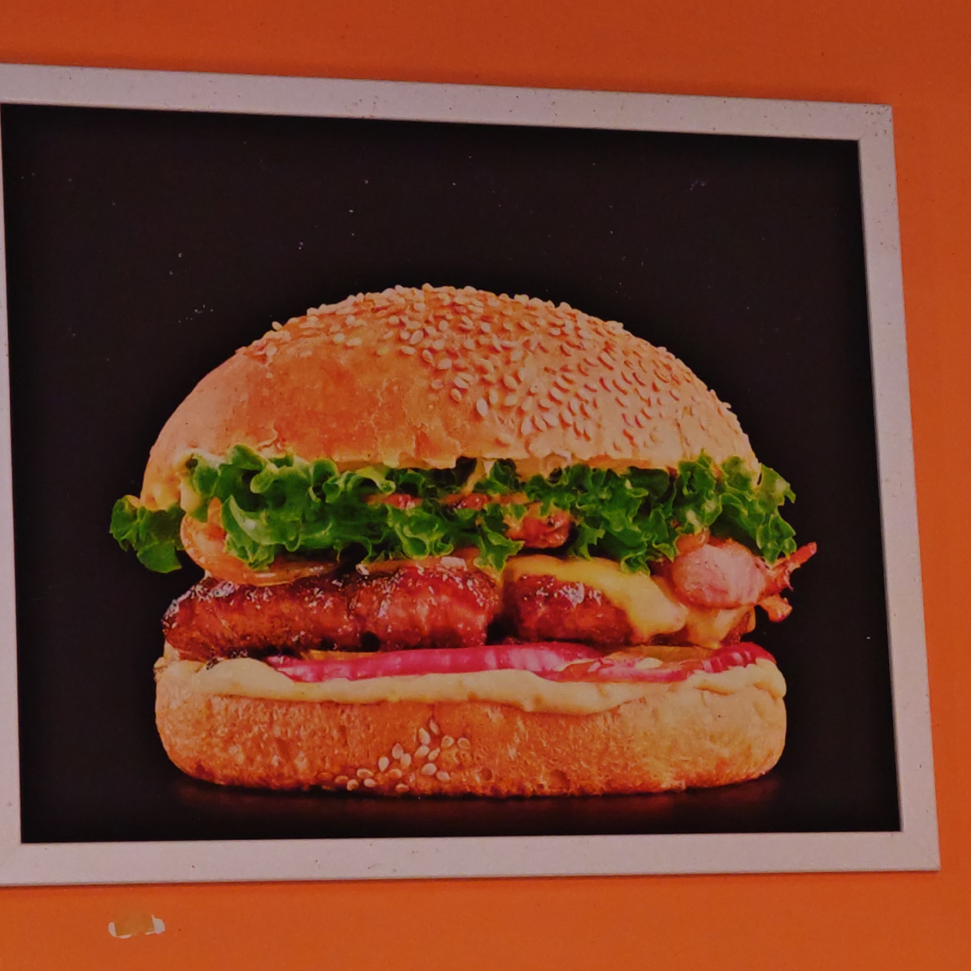 The image is a framed photograph of a large hamburger with sesame seeds on the bun, lettuce, meat, cheese, and other toppings, set against a black background. The frame is white and the wall behind it is painted orange. The location is the city of Rostov, Russia.