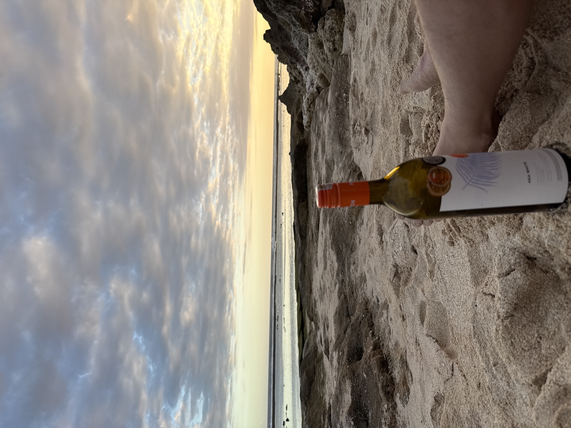 The image shows a beach at Kuta Selatan, Indonesia, with a bottle of Aza White wine lying on the sand next to a person's feet. The sand is light brown and textured. In the background, the ocean meets a cloudy sky with a hint of sunset. Rocky cliffs are visible on the right side of the image.