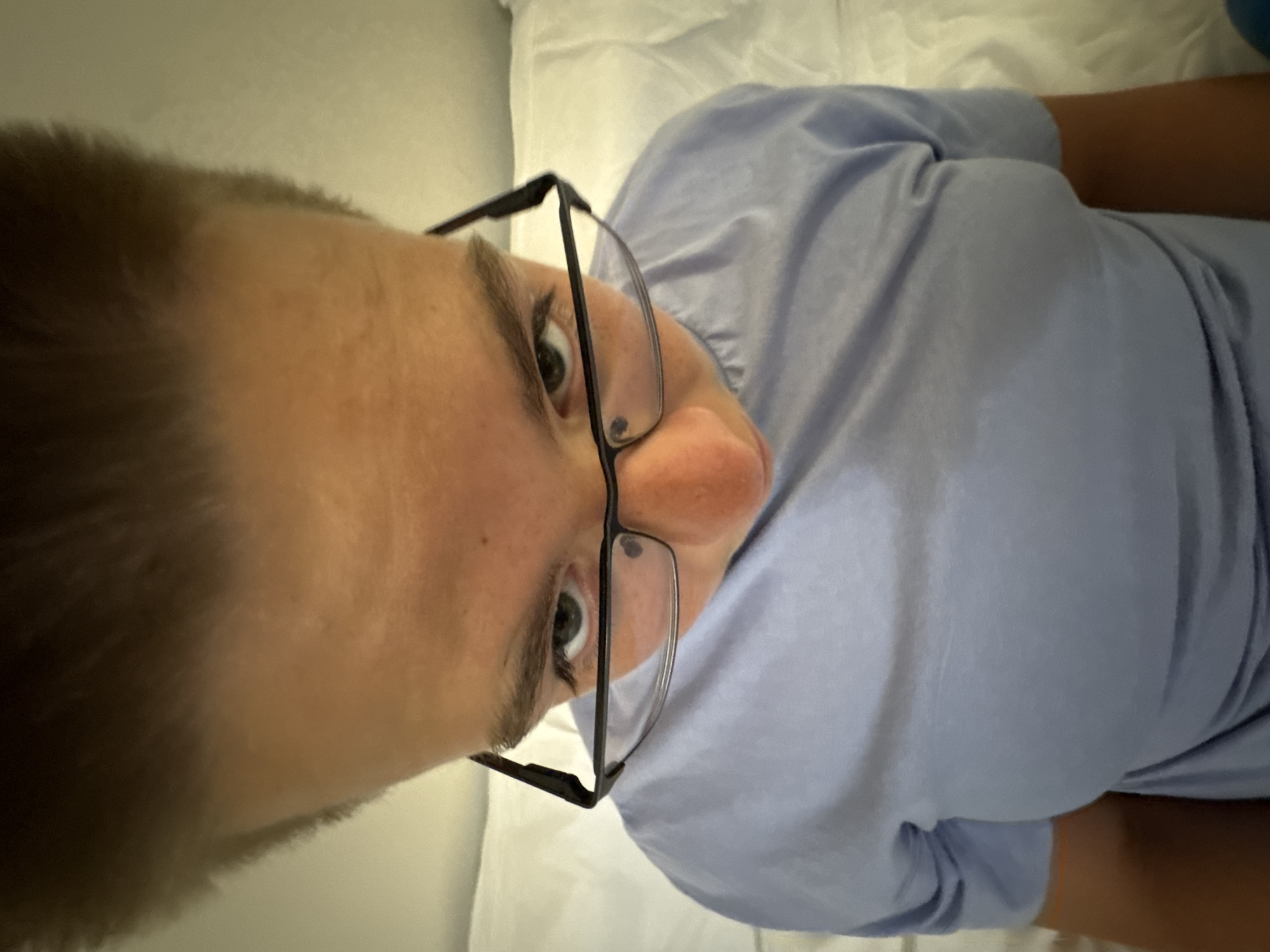 A close-up, high-angle shot shows a young person wearing glasses and a light blue t-shirt, lying down on a white, textured surface that appears to be a bed or couch. The person's face is the central focus, with their eyes looking directly at the camera. The lighting is soft, casting subtle shadows. The background is blurred, with the white surface dominating. The image does not provide any information about the city of Nesebar, Bulgaria, as there are no discernible landmarks or contextual clues to place it geographically.