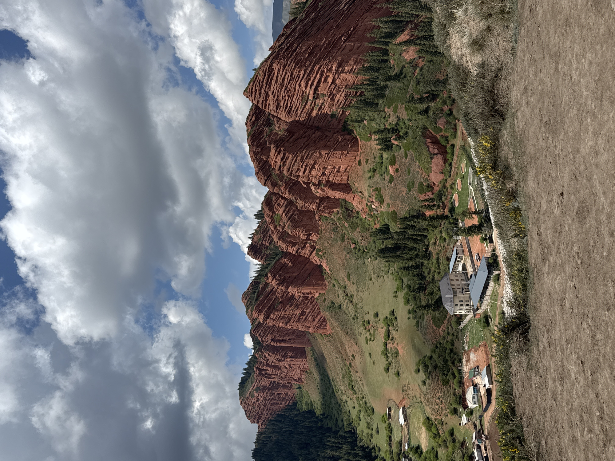 This media file captures a wide, scenic view of the Jeti-Ögüz gorge in Kyrgyzstan. The primary subjects are the striking red rock formations that stretch across the left and central parts of the image. These massive, layered cliffs, characteristic of Jeti-Ögüz, feature deep striations and varying shades of reddish-brown, contrasting with patches of dark green pine trees and other vegetation growing on their slopes and at their bases.
In the middle ground, nestled at the foot of these geological features and surrounded by green, grassy hills, is a collection of buildings. A prominent multi-story structure, possibly a hotel or resort, with a light-colored exterior and darker roofs, is visible. Several smaller buildings and outbuildings are also present, some resembling houses or support facilities. Towards the left of these structures, a few circular, manicured green areas suggest a golf course or recreational space. Further in the background, closer to the base of the red rocks, more distant, smaller structures or yurts can be observed.
The sky above is partially cloudy, showcasing a mix of bright blue patches and large, white to grey cumulus clouds. The foreground on the right consists of dry, earthy terrain, possibly a hillside or a dirt track, with patches of dry grass. A distinct line of small yellow flowers grows along what appears to be a fence or boundary separating the drier foreground from the greener developed area.
The image depicts a tranquil, daytime scene, likely under fair weather conditions, given the bright light and scattered clouds. No specific human activity is visible, but the presence of buildings indicates human settlement or a tourist destination within this rugged natural landscape. There is no visible text in the image.