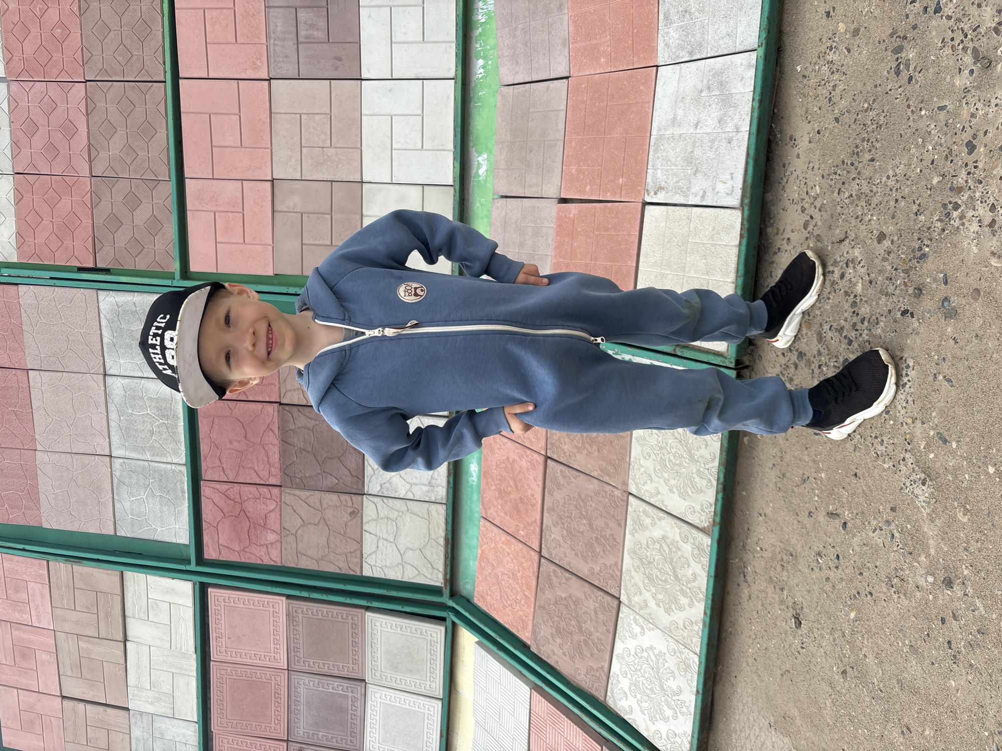 A young boy stands in front of a display of paving tiles. He wears a blue jumpsuit, black sneakers with white soles, and a black and white baseball cap with the word "ATHLETIC" on it. The tiles behind him are arranged in a variety of colors and patterns, framed by a green metal structure. The ground is concrete. The image was taken outdoors in Chaikovsky, Russia.