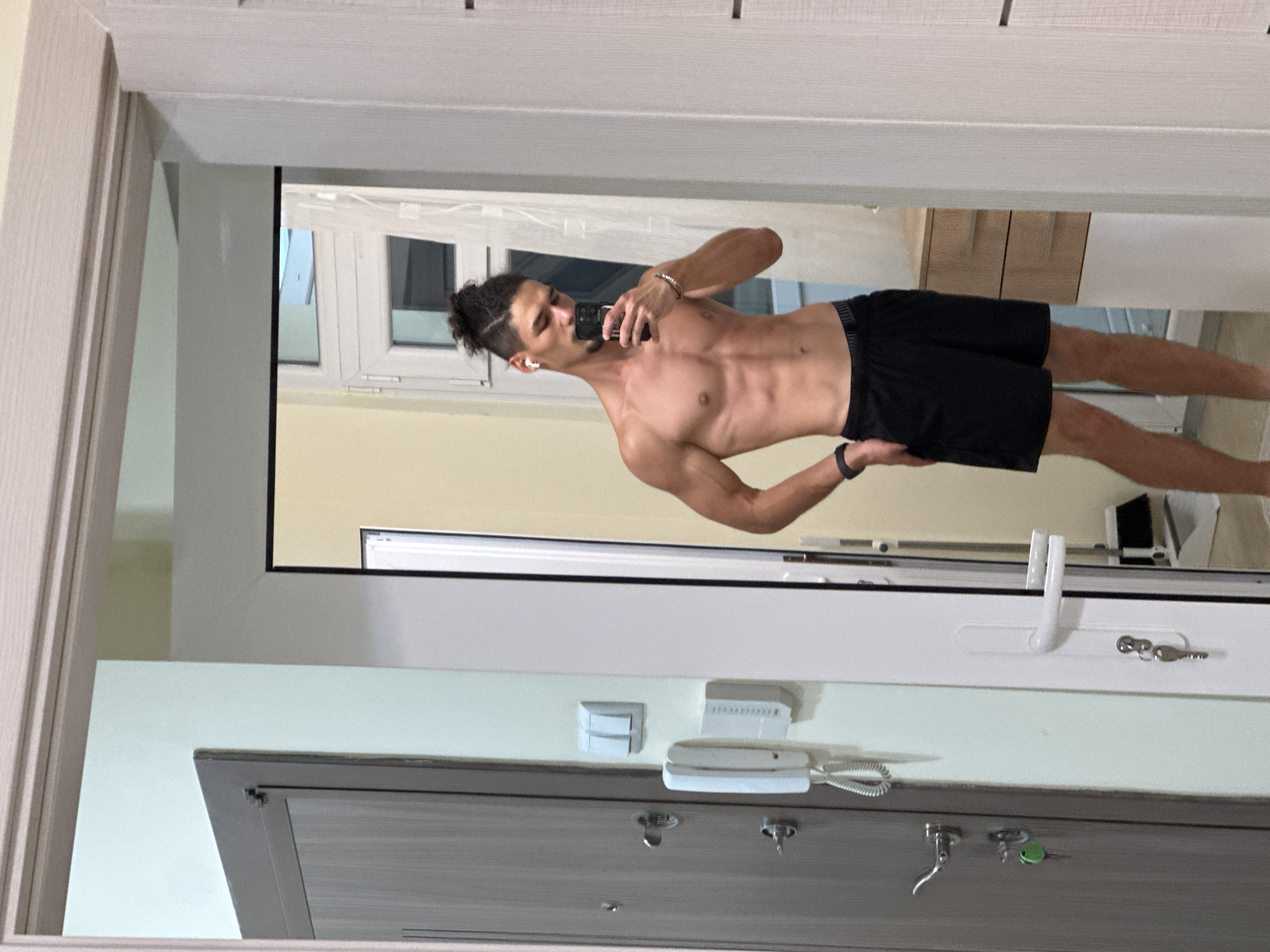 A shirtless man with dark hair pulled into a bun and a light beard is taking a mirror selfie in a hallway. He is wearing black shorts and wireless earbuds. His left hand is on his hip, and he is holding his phone in his right hand. The hallway has a light-colored wall, a gray door on the left with a white intercom and light switches, and a white door frame on the right. In the reflection, a window with white blinds is visible, as is a wooden dresser with two drawers. The image is a close-up of the man and the immediate surroundings.