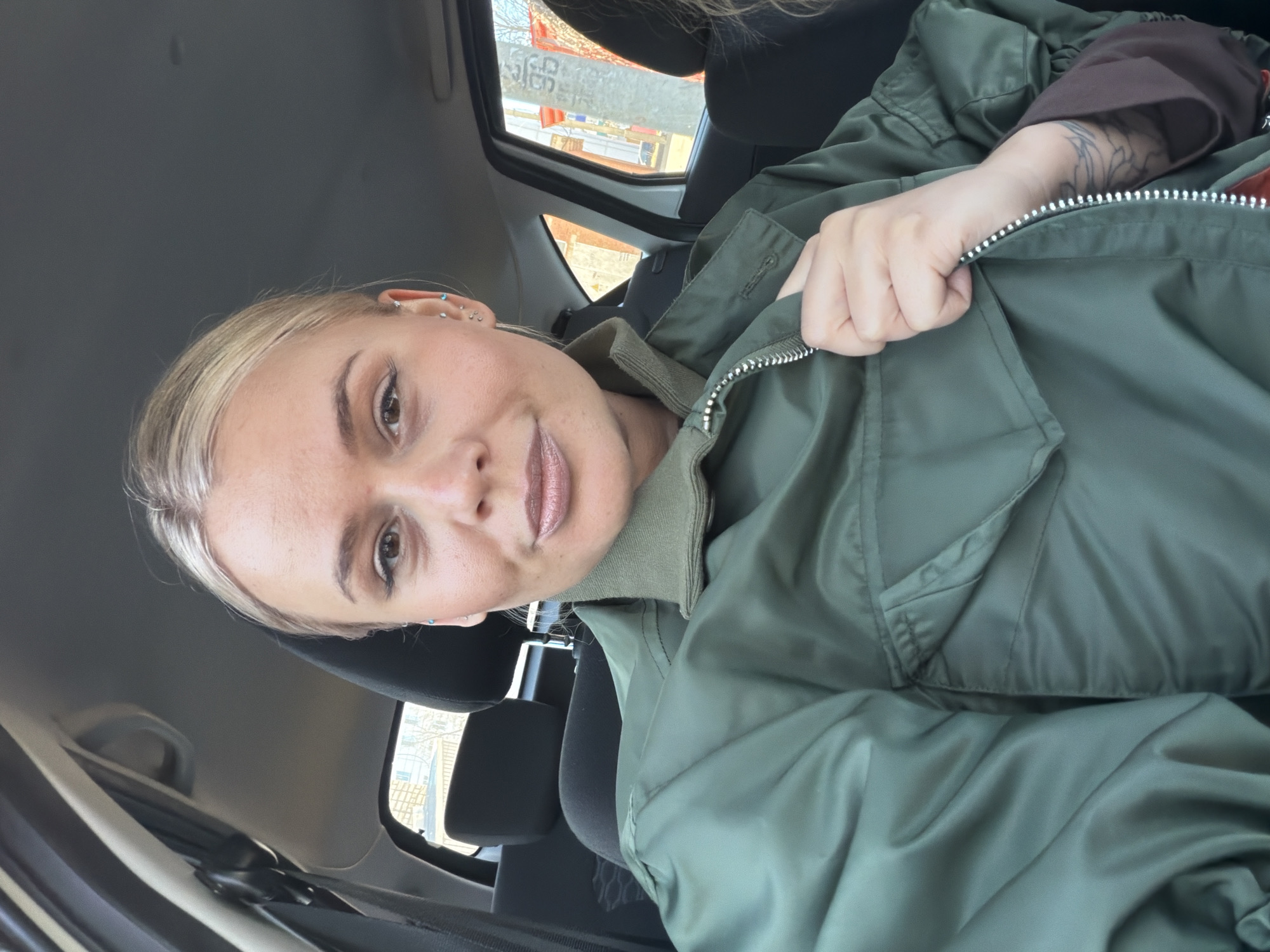 A woman is seated in a car. She has blonde hair and multiple ear piercings. She is wearing a green bomber jacket and appears to be adjusting the zipper. The environment visible through the car windows shows an urban setting with buildings and signs, possibly the city of Sarapul, Russia, though this is unconfirmed by the image alone.