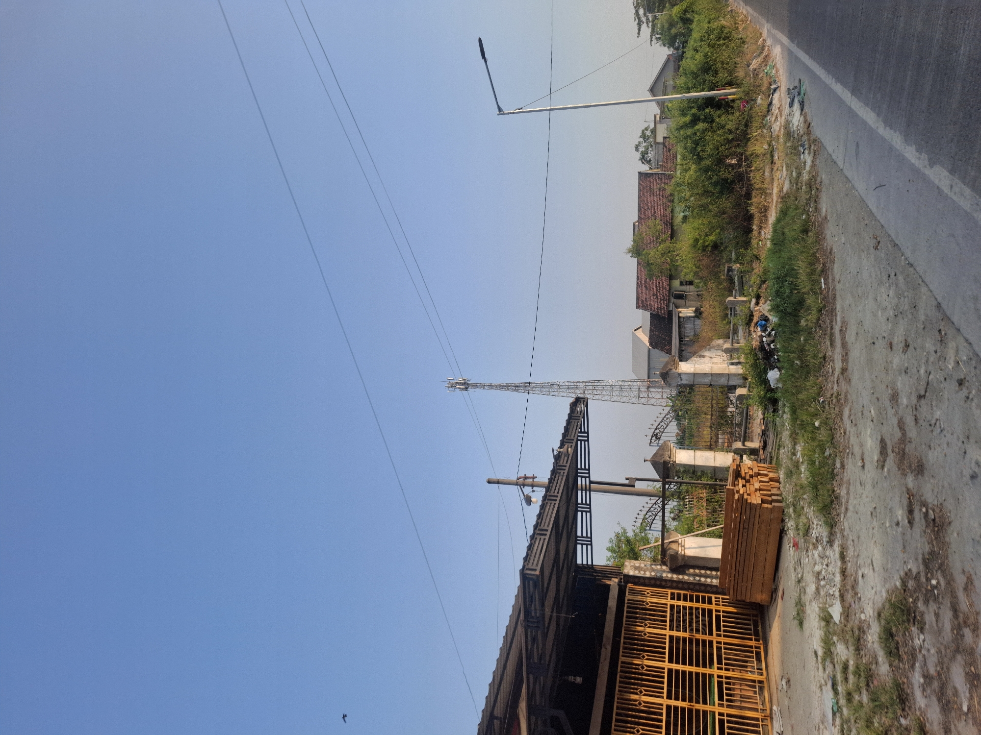 The image captures an outdoor scene during daylight in what appears to be a rural or semi-urban area of Sungelebak, Indonesia. The sky is clear and blue, with a few faint power lines stretching across it. In the foreground, a paved road runs along the right side of the frame. To the left of the road, there is a dirt and gravel area with sparse grass and some scattered debris.
Adjacent to this area is a structure with a dark, corrugated metal roof supported by a metal frame, extending over a bright yellow metal gate. Stacked neatly to the left of the gate are several wooden planks. Behind the gate and planks, a weathered stone pillar and a section of a fence are visible. Further into the background, a tall telecommunications tower stands prominently. Beyond the tower, a cluster of houses with red tiled roofs are nestled amongst lush green trees and bushes. A street lamp is positioned on the right side of the road, and a small bird is seen in flight in the upper left portion of the sky. The overall impression is one of a functional, perhaps commercial, area bordering a residential and natural landscape.