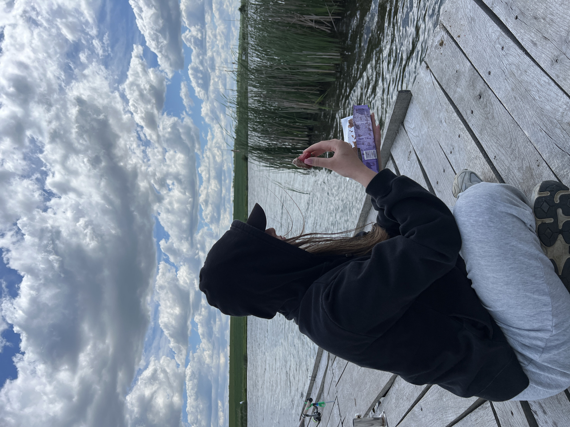 A person wearing a black hooded sweatshirt and gray sweatpants sits on a wooden dock, holding a small piece of food, possibly a strawberry, in their right hand and a purple snack wrapper in their left hand. The dock extends over water with tall reeds growing along the shoreline in the background. Fishing rods are visible at the edge of the dock near the person's feet. The sky is partly cloudy with blue patches.
The location is not identifiable as Kopychyntsi, Ukraine. The scene depicts a person on a dock, potentially fishing and eating. The environment includes water, reeds, a wooden dock, and a partly cloudy sky.