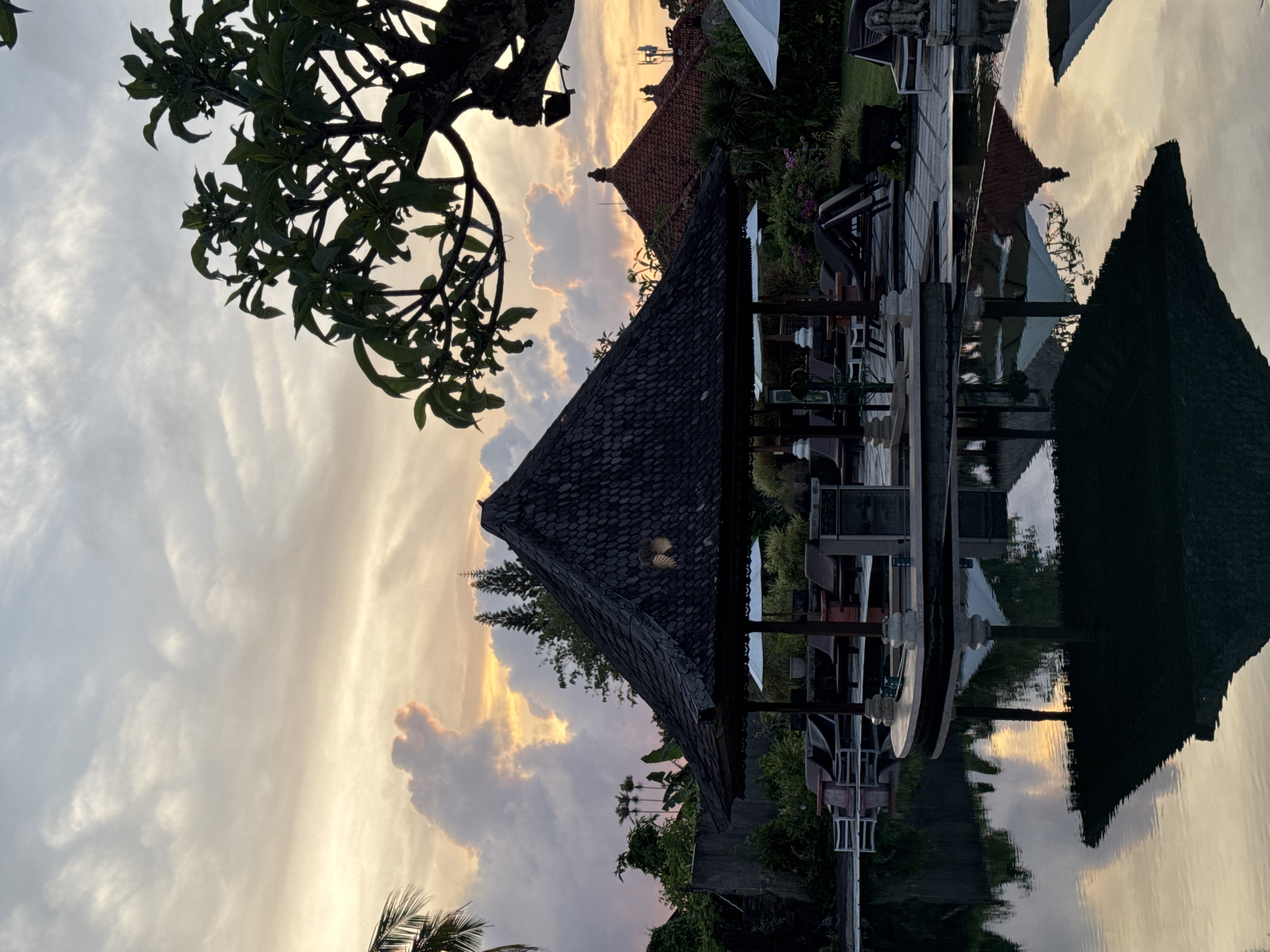 The image depicts a serene scene, likely at sunset, in Jimbaran, Indonesia. The focal point is a traditional Balinese pavilion or gazebo with a dark, tiled roof, positioned near a body of water that reflects the sky and surrounding structures, creating a mirror effect.
The sky transitions from shades of light blue to golden hues, indicating either sunrise or sunset. Clouds are present, adding depth and texture to the sky.
Vegetation, including trees and tropical plants, surrounds the pavilion. The branches of a tree with dense foliage are seen in the upper left corner. Additional buildings with reddish roofs are visible in the background, implying a resort or residential area.
The calm body of water reflects the pavilion, trees, sky, and buildings. Outdoor seating and dining areas with tables, chairs, and possibly other amenities are suggested by the structures adjacent to the water. The scene is peaceful and evocative of a tropical setting.