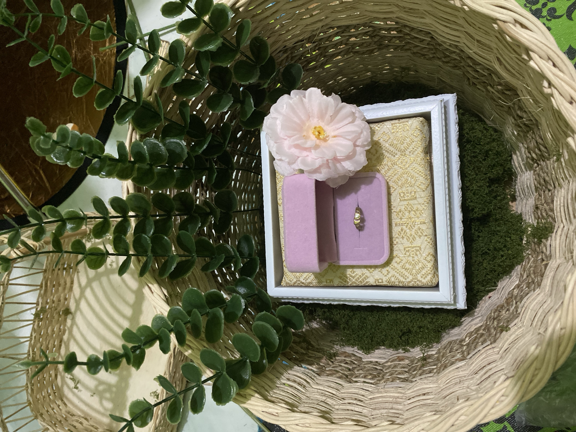 This image captures a close-up view of a decorative arrangement within a woven basket, likely for a special occasion such as a proposal or wedding. The centerpiece is a small, white-framed display holding a ring. The ring, appearing to be gold and set with small stones, rests within a pink velvet ring box. To the left of the ring box, a delicate pale pink fabric flower with a yellow center is placed, adding a touch of softness and color. The frame is set upon a textured gold fabric, which in turn sits on a bed of green moss.
The basket itself is made of natural, light-colored woven material, creating a rustic and earthy feel. To the left of the basket, artificial green foliage with small, round leaves cascades down, framing the scene and adding a natural element. The lighting suggests an indoor setting, possibly with soft, ambient light. The overall presentation is one of careful curation and romantic sentiment, hinting at a significant personal event. There is no visible text in the image. The location context of Kuala Pembuang, Indonesia, suggests this could be part of a traditional ceremony or celebration in that region.