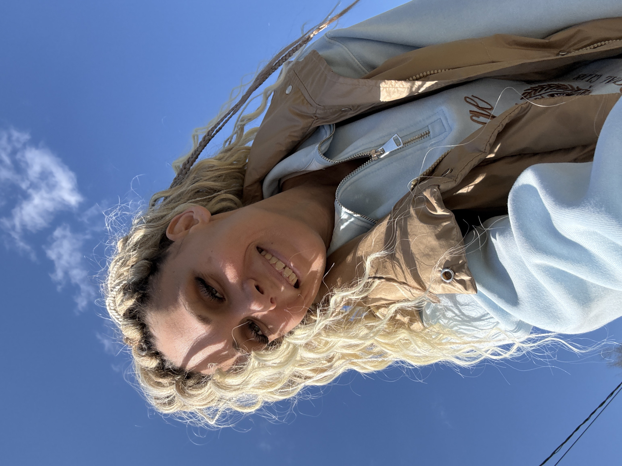 A woman with blonde, braided hair smiles into the camera, framed by a clear blue sky with scattered clouds. She wears a light blue zippered hoodie under a tan jacket. The location is unknown; there is no evidence to indicate the location is Rusanovo, Russia.