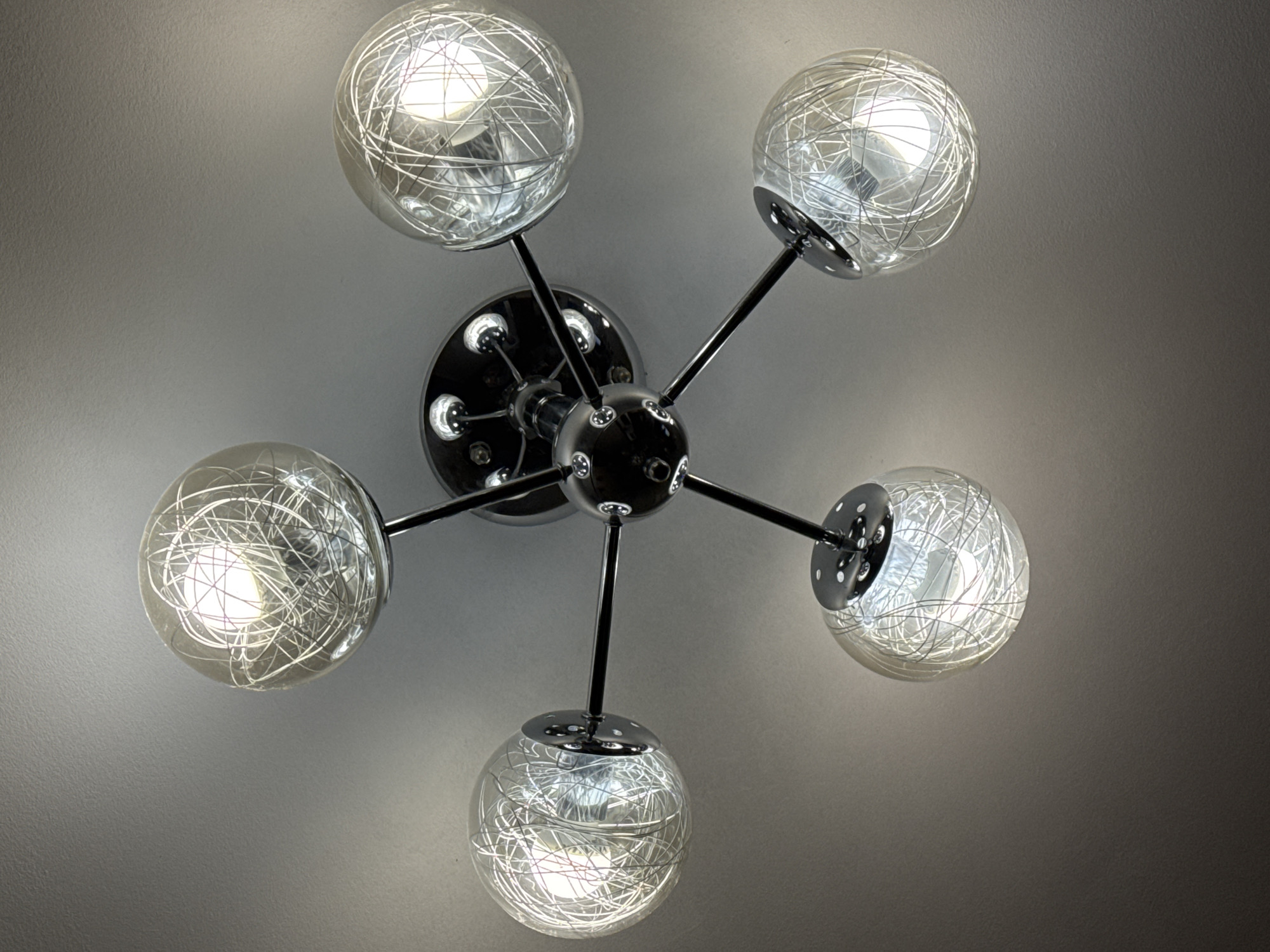 The image shows a close-up, low-angle shot of a modern, five-light ceiling fixture. The fixture has a chrome-plated base with a central disc from which five arms extend outwards. Each arm terminates in a spherical glass shade that contains a tangled wire or filament design. The bulbs within the shades are illuminated, casting a soft light. The fixture is mounted on a grey, textured ceiling. The image does not contain any information about the city of Burgas, Bulgaria, or any people, actions, or interactions.