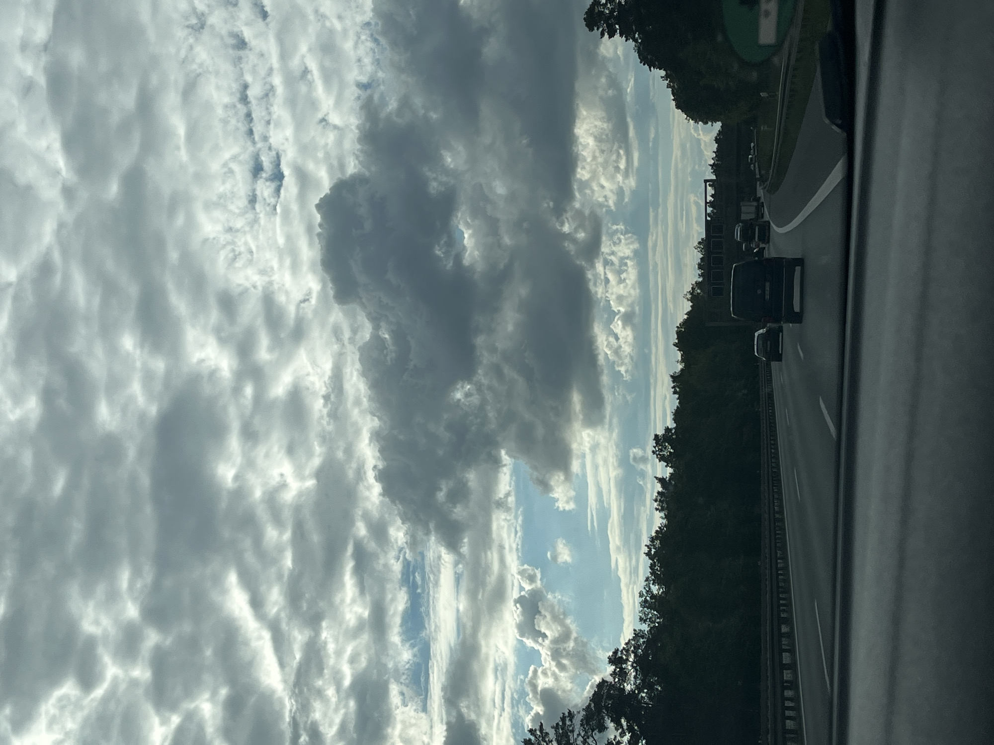 The image shows a view from inside a vehicle traveling on a highway. The sky is filled with clouds, predominantly grey and white, with some patches of blue visible. Adjacent to the highway is a line of trees. Several vehicles, including a dark-colored van, are seen on the road ahead. The perspective suggests the photograph was taken from the passenger seat. The exact location is unknown.