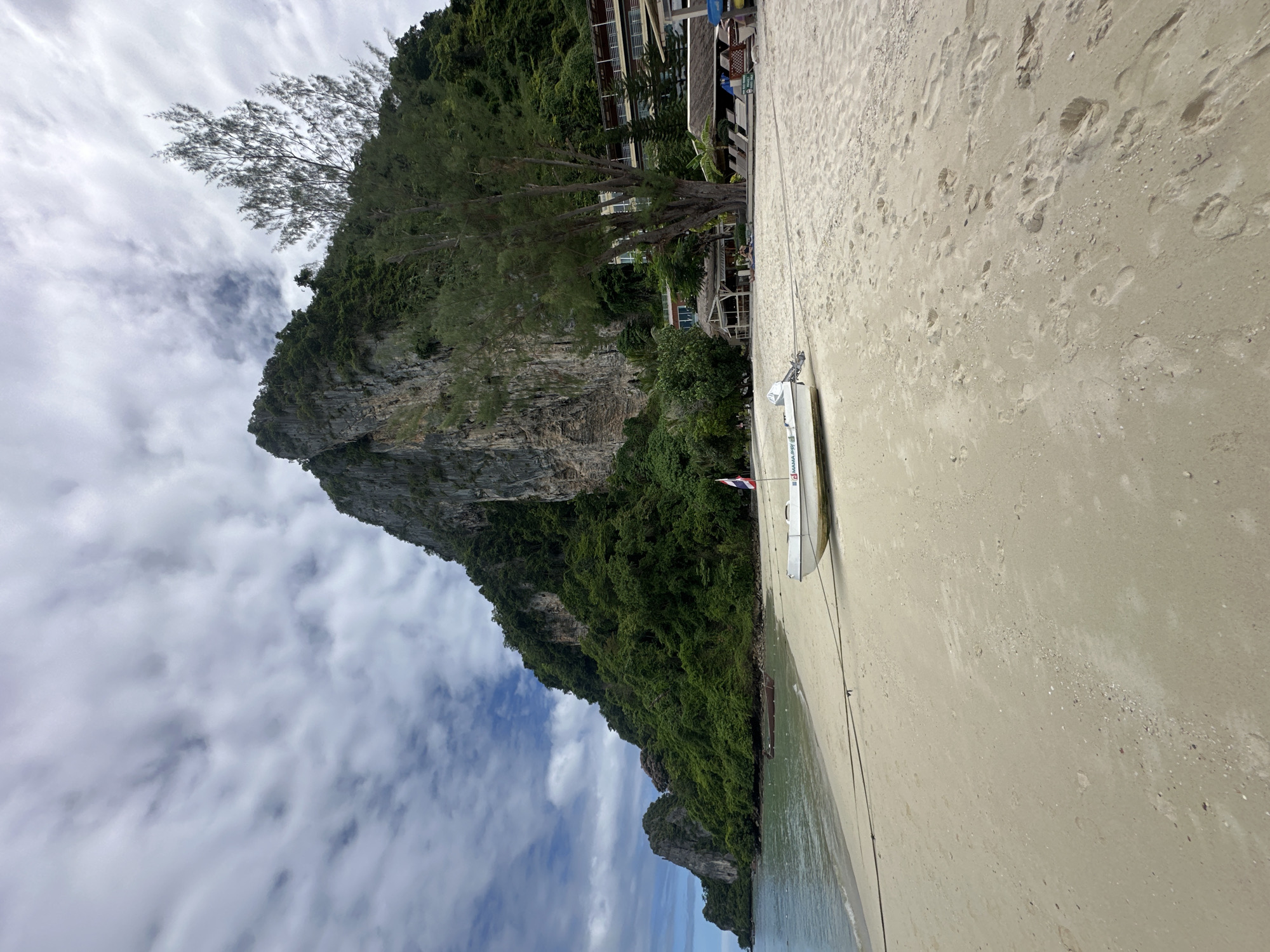 A white, inflatable boat with a small outboard motor and a Thai flag is pulled up onto a sandy beach in Ao Nang, Thailand. The boat is tethered by a rope that stretches across the sand to the left. In the background, lush green vegetation covers a steep, rocky cliff face. The sea is calm and a few other small boats are visible in the distance. Several resort buildings with balconies are nestled into the hillside behind the beach. The sky is partly cloudy with dramatic, wispy formations. Footprints are visible in the sand, suggesting recent activity.