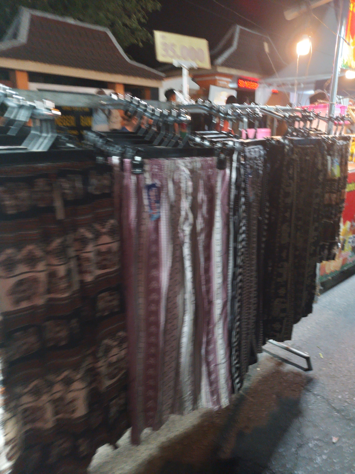This image depicts a street market stall at night in Kertosono, Indonesia. The stall displays a variety of textiles, including patterned fabrics with what appear to be motifs of elephants and geometric designs, hanging from metal racks. Some of the fabrics are draped vertically, with distinct stripes of white, pink, and grey visible. The background shows the roof of a building with traditional architecture, illuminated by artificial lights, and a bright yellow sign with the number "35.000". A street lamp is also visible, casting light on the scene. The ground appears to be asphalt or concrete. The overall impression is a bustling night market environment.