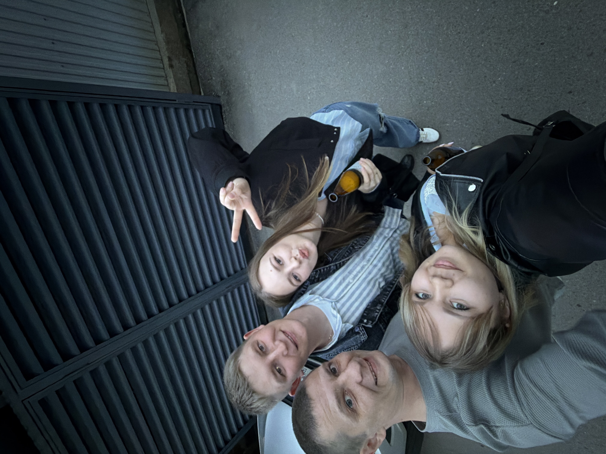 A low-angle, wide-shot selfie captures four individuals in the city of Chaikovsky, Russia. From left to right, a male with short dark hair in a grey long-sleeved shirt smiles at the camera. To his right, a blonde-haired male in a striped light blue and white shirt and denim jacket also smiles. A blonde-haired female rests her head on the first male's shoulder, smiling at the camera, with a partially visible brown bottle in her right hand. Above and to the right of the blonde male, another female with long brown hair poses, making a peace sign with her right hand and holding a brown bottle in her left. She wears a black jacket, light blue shirt, and ripped jeans, and makes a puckered lip expression. The background features a dark grey slatted metal structure, possibly a gate or facade, and a lighter grey corrugated metal wall to the right. A white vehicle is partially visible on the far left. The ground is dark grey asphalt.
