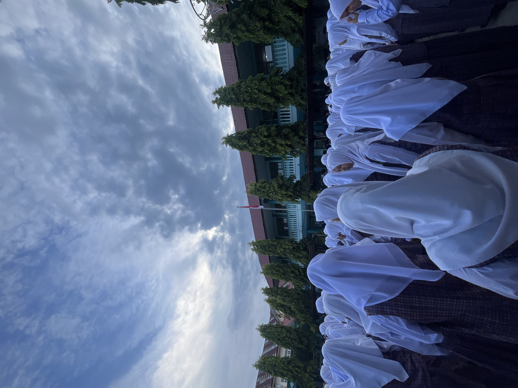 An outdoor, low-angle medium shot captures a group of school-aged females, identifiable by their white headscarves and plaid skirts, gathered in what appears to be a schoolyard in Lhokseumawe, Indonesia. The individuals are predominantly facing away from the camera, their heads tilted upwards towards a cloudy, overcast sky. In the background, a two-story building with a red roof and teal-colored balconies is visible, lined with tall, conical green trees. A flagpole with the Indonesian flag is positioned to the right of the building. The overall atmosphere suggests a school assembly or ceremony.