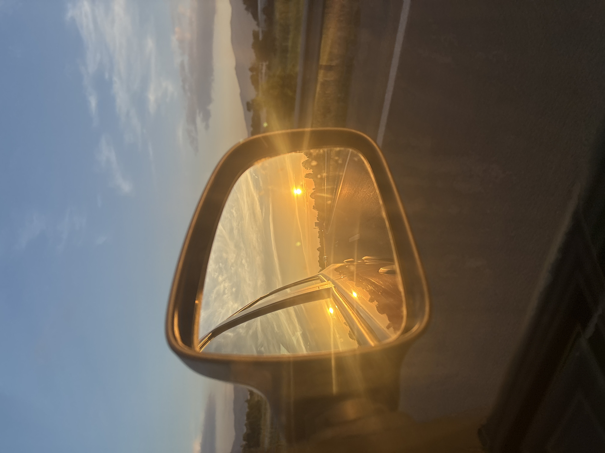 The image shows a view of a sunset reflected in a car's side mirror. The reflection reveals a road stretching into the distance, with a bright sun low on the horizon. Wispy clouds streak across a pale blue sky, tinged with orange and yellow from the setting sun. The foreground of the image, outside the mirror, shows the car's interior and the road it is traveling on. There are no people visible in the image. The location in the city of Sofia, Bulgaria, is not discernible from the visual information provided.