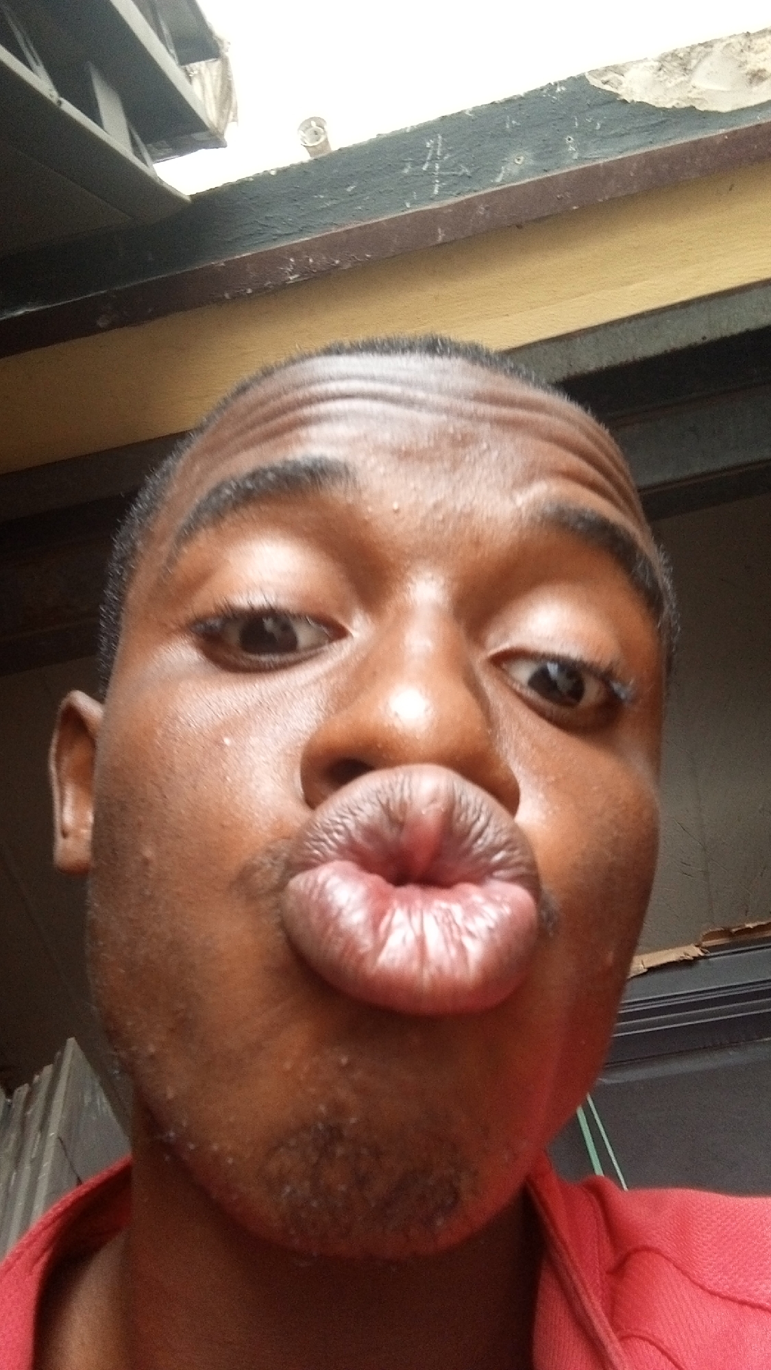 A close-up selfie of a Black male with dark brown eyes and short black hair, pursing his lips in a kissing gesture. The background consists of a building exterior with visible roof beams and a partial view of the sky. The person is wearing a red shirt. The location is identified as Nkpor, Nigeria.