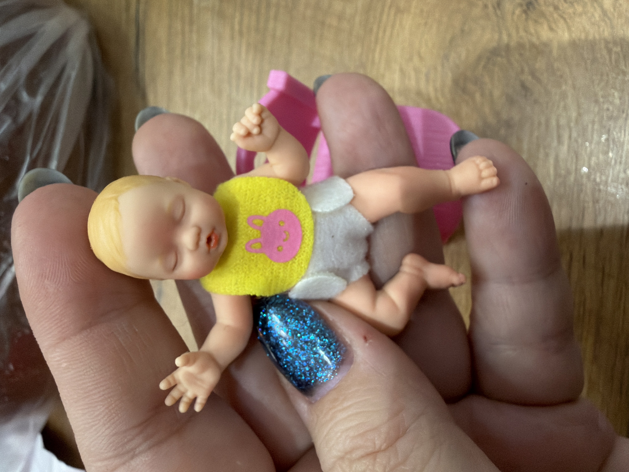 A small, plastic baby doll is held in the palm of a person's hand. The doll has blond hair, closed eyes, and a yellow bib with a pink bunny design. The doll wears a white diaper. The hand holding the doll has short nails, one of which is painted with a glittery blue polish. A pink plastic object, possibly part of a toy or accessory, sits behind the doll on the hand. The background is a blurred brown surface with a hint of a plastic bag in the upper left corner. There is no evidence indicating this image was taken in Chaikovsky, Russia.