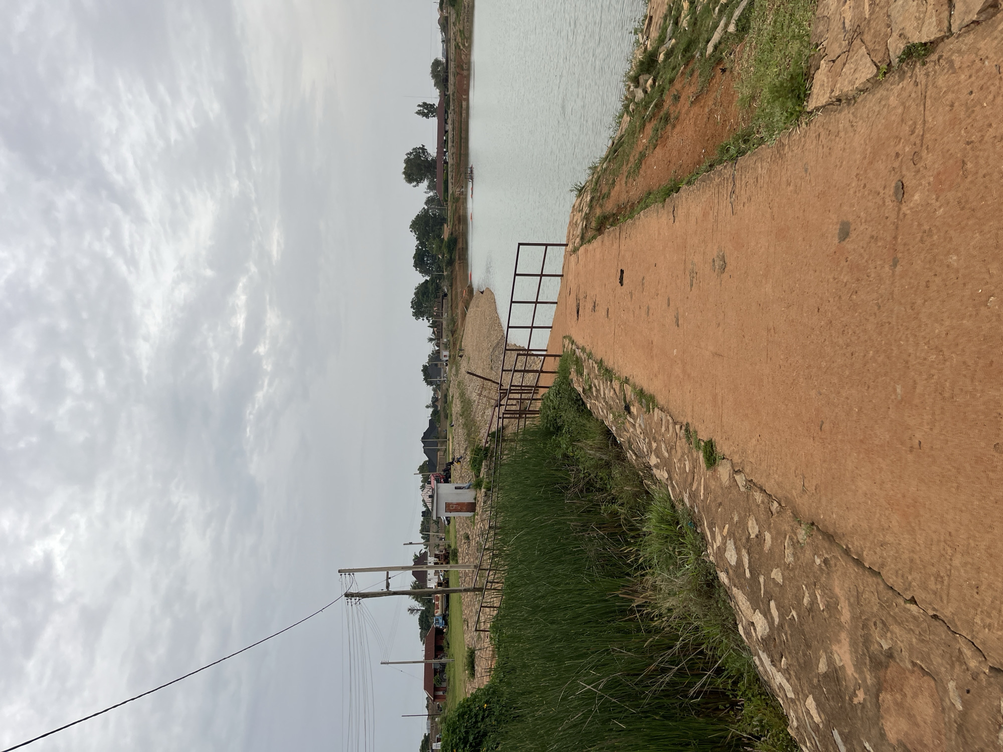 The image depicts a tranquil scene at a reservoir or lake in Bukuru, Nigeria. A paved pathway, made of reddish-brown concrete, leads towards the water. The pathway is bordered by a metal railing on the left, which runs along the edge of the reservoir. Tall green reeds and grasses grow along the left side of the pathway, separated by a rocky embankment. The reservoir itself is a calm body of water, reflecting the overcast sky. In the background, several buildings, including what appears to be a small white structure, a red-roofed building, and other residential or industrial structures, are visible. Utility poles and power lines are also present. A truck and a tractor are parked near the buildings, suggesting some ongoing activity. The overall atmosphere is peaceful, with the muted colors of the sky and landscape contributing to a serene ambiance.