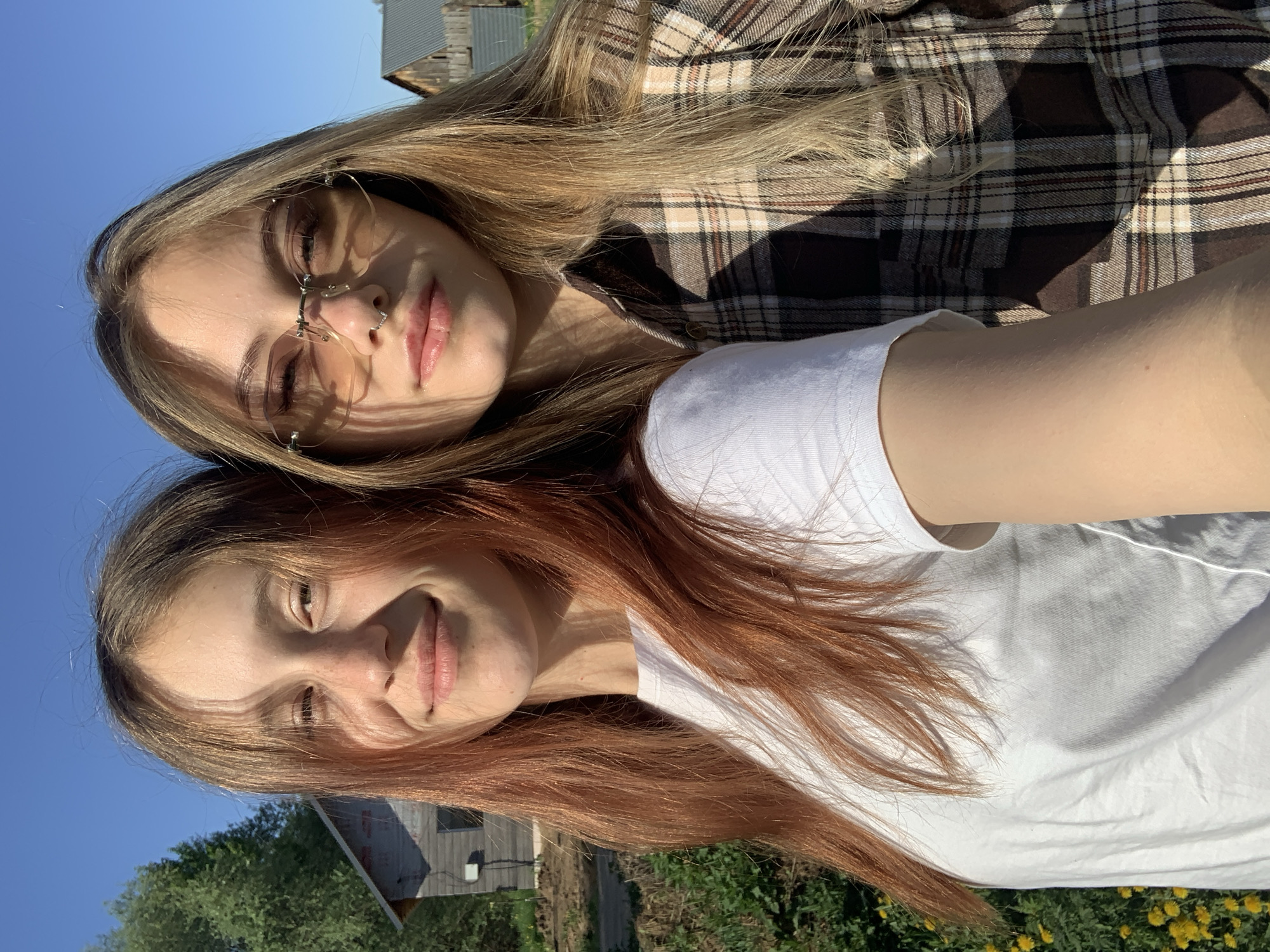 Two young women are lying next to each other outdoors. The woman on top has blonde hair and wears glasses and a brown plaid shirt. The woman below has red hair and wears a white t-shirt. The background shows grass, dandelions, and some buildings. The location may be in Фоки, Russia.