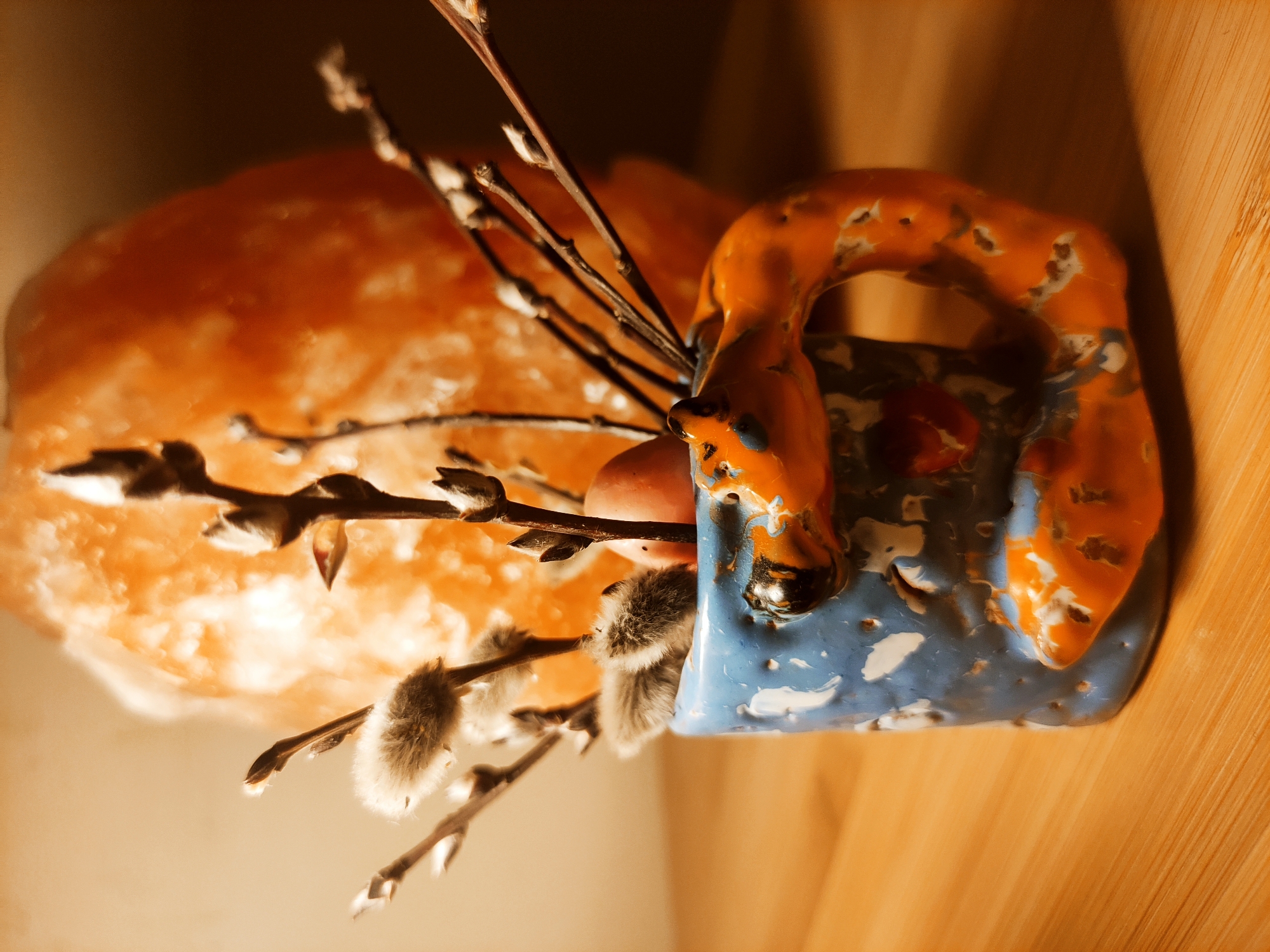 Here's a description of the image:
The image depicts a still life arrangement. A rough, orange-colored salt lamp is positioned to the left of a small, hand-painted ceramic cup. The cup is primarily blue and orange with white accents and an irregular shape. Several sprigs of pussy willow are arranged within the cup and against the lamp. The arrangement rests on a wooden surface. The lighting is warm, creating shadows and emphasizing textures. The scene appears to be indoors. The city and exact location are not identifiable from the image.
