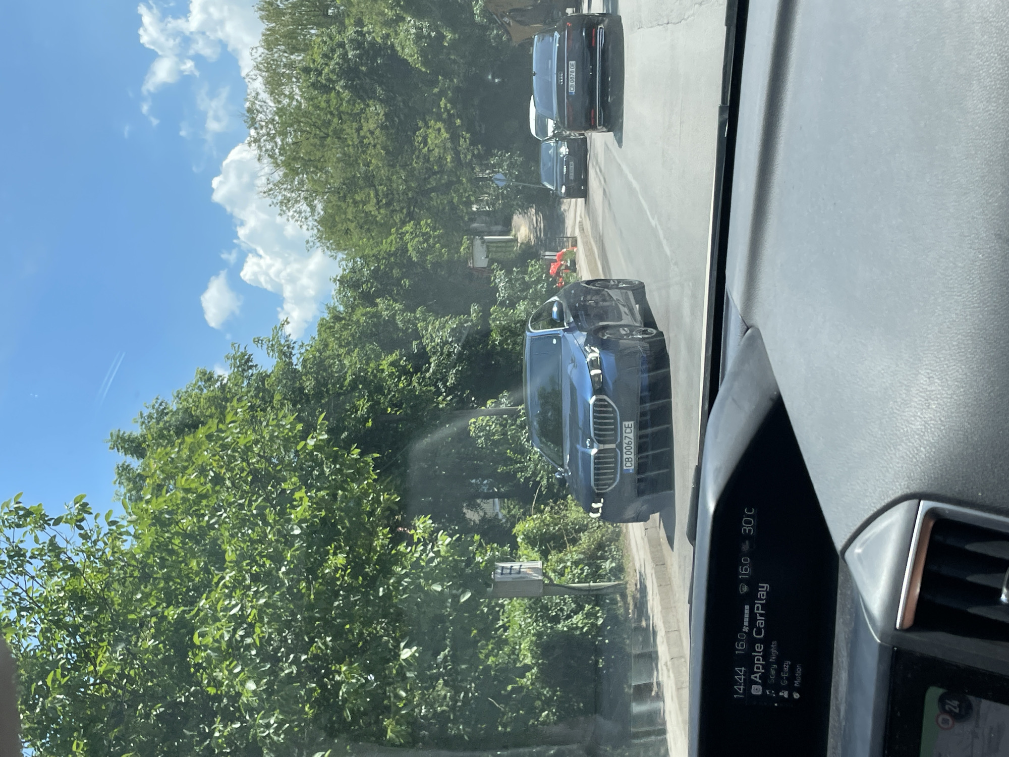 The image depicts a street scene in Sofia, Bulgaria, viewed from the interior of a vehicle. The foreground shows the dashboard of the vehicle, with the display screen of "Apple CarPlay" visible, showing the time as 14:44, a temperature of 16.0°C, and another temperature reading of 30°C.
Outside the vehicle, a blue BMW sedan with license plate "CB 0067 CE" is prominent in the lane. Further down the road, an Audi sedan with license plate "CB 8878 OE" is visible, along with a white vehicle and another dark sedan. The road is lined with dense green trees and foliage on both sides, with a sidewalk visible on the left. The sky is blue with scattered white clouds. A traffic sign is partially visible on the left side of the road, and construction or road work equipment can be seen in the distance on the right. The overall impression is of a sunny day in a tree-lined urban or suburban environment in Sofia.