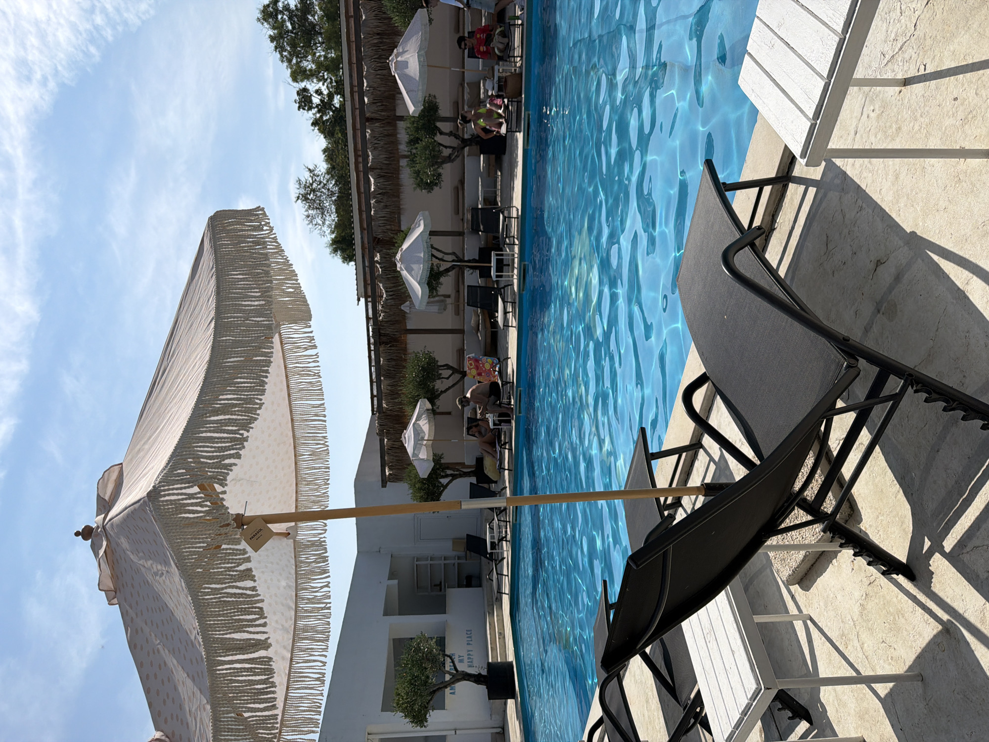 A high-angle, full shot captures a poolside scene on a sunny day. In the foreground, two black lounge chairs with small white tables are positioned on a light-colored concrete patio. A large, cream-colored umbrella with decorative fringe is prominently displayed, partially obscuring the sky. Beyond the chairs, a bright blue swimming pool occupies the center of the frame.
In the middle ground, several people are visible relaxing on lounge chairs near the pool. To the left, a white building with an open-air reception area and an olive tree is visible, with text on the wall reading "AM BEACH MY HAPPY PLACE". Further back, more lounge chairs and white umbrellas are arranged, and trees with thatched roofing create a shaded area. A few more individuals are seen lounging in this area.
The background consists of more trees and the sky, which is a vibrant blue with scattered white clouds. The overall impression is a resort or hotel setting designed for relaxation and leisure. The scene appears to be located at the AM Beach located in the city of Burgas, Bulgaria, given the prominent text on the building.