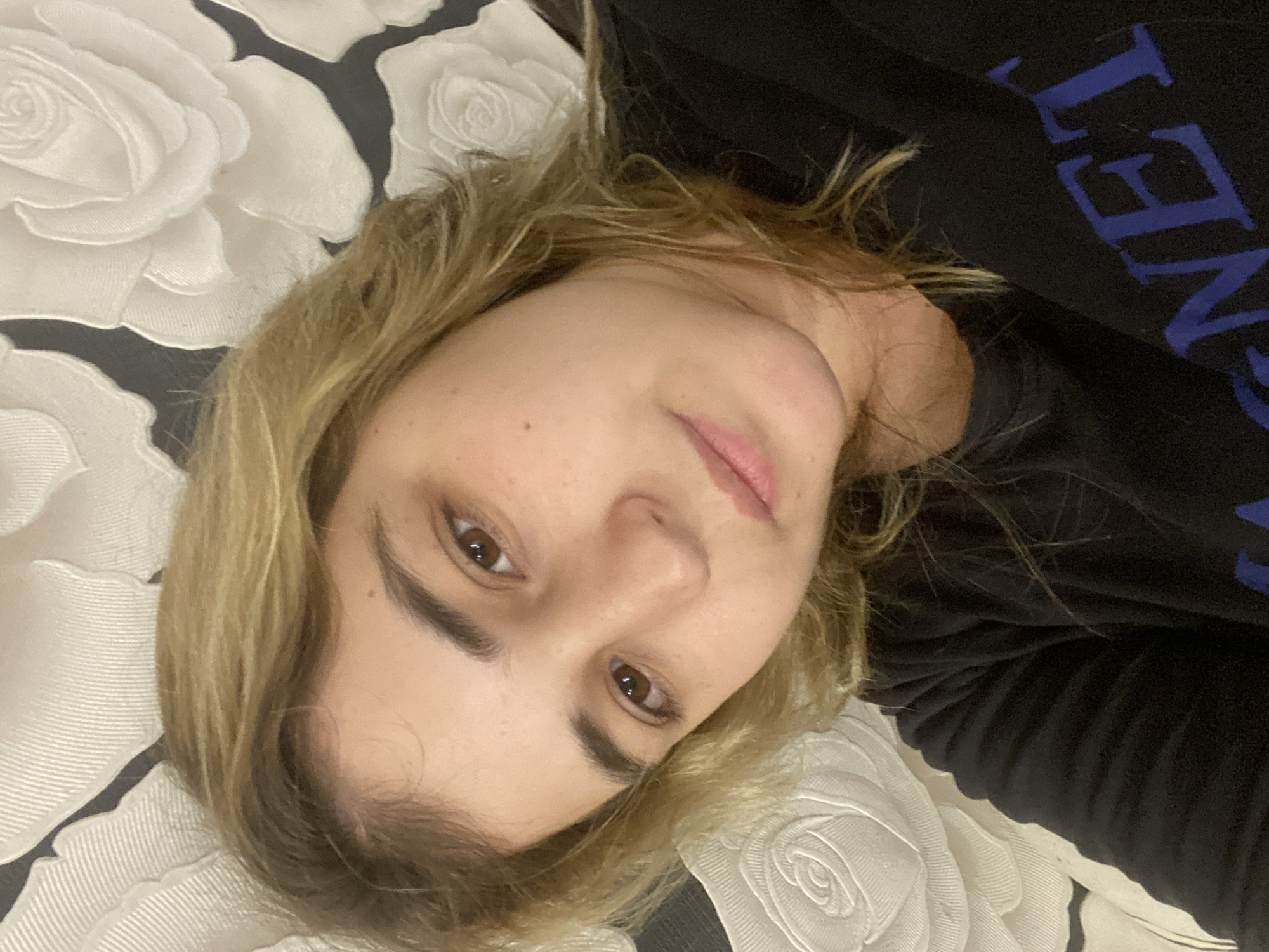 A young woman with blonde hair and brown eyes is lying down, looking at the camera. She is wearing a black sweatshirt with blue lettering on it. The background is a textured wallpaper with a white floral pattern. The image is a selfie, taken from a close-up angle. The lighting is soft and natural. There are no other people or objects in the image. The location is not specified in the image, but the prompt states it is Chegem-Vtoroy, Russia.