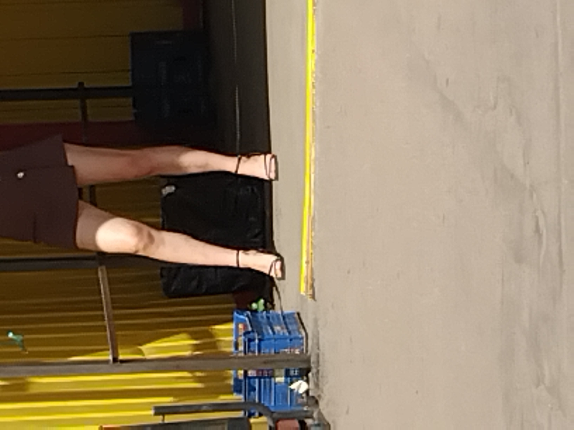 The image captures a segment of a person's legs, from mid-thigh down to the feet, dressed in a dark skirt and black stiletto-heeled sandals. The individual is standing on a paved surface with a yellow line marking a boundary. In the background, a yellow corrugated metal wall is visible, along with stacks of blue plastic crates to the left and a black object, possibly a large bag or a piece of equipment, behind the person. The overall setting appears to be an outdoor urban environment, possibly a loading dock or a service area within the city of Zhytomyr, Ukraine, given the context provided. The lighting suggests it is daytime.
