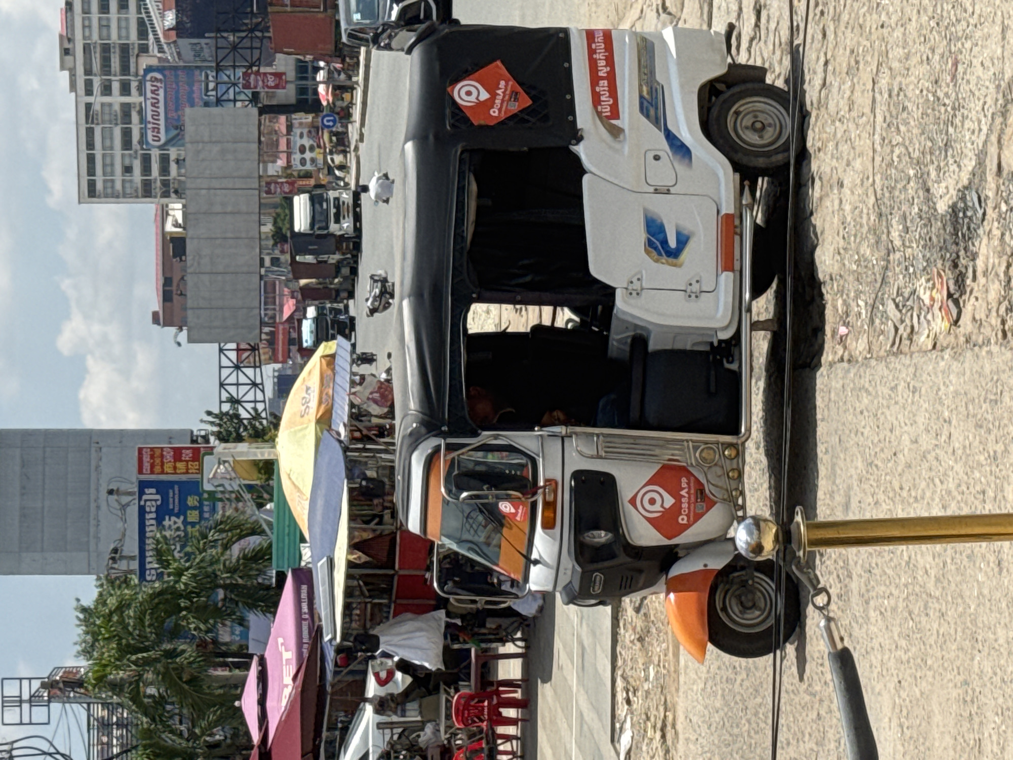 The image captures a street scene in Bavet Quarter, Cambodia, on a bright, possibly hot, day with a partly cloudy sky. In the foreground, a white and orange tuk-tuk, a common mode of transport in Southeast Asia, is prominently parked on a cracked, dusty road. The tuk-tuk features a black roof and tinted windows, and it displays the "PassApp" logo in orange and white on its side. The road is lined with cables and a golden bollard.
Behind the tuk-tuk, a bustling market area is visible. Several market stalls are set up with colorful umbrellas, including pink, red, and yellow ones. These stalls appear to be selling various goods, with items like clothing and bags visible. Red plastic chairs are stacked near one of the stalls, suggesting a seating area. In the background, tall buildings with modern architecture stand out, some appearing to be commercial or residential structures. Several trucks and other vehicles are on the road, indicating traffic. A few individuals are seen in the distance, including someone riding a motorcycle. The overall impression is of a busy, dynamic urban environment typical of a Cambodian city.