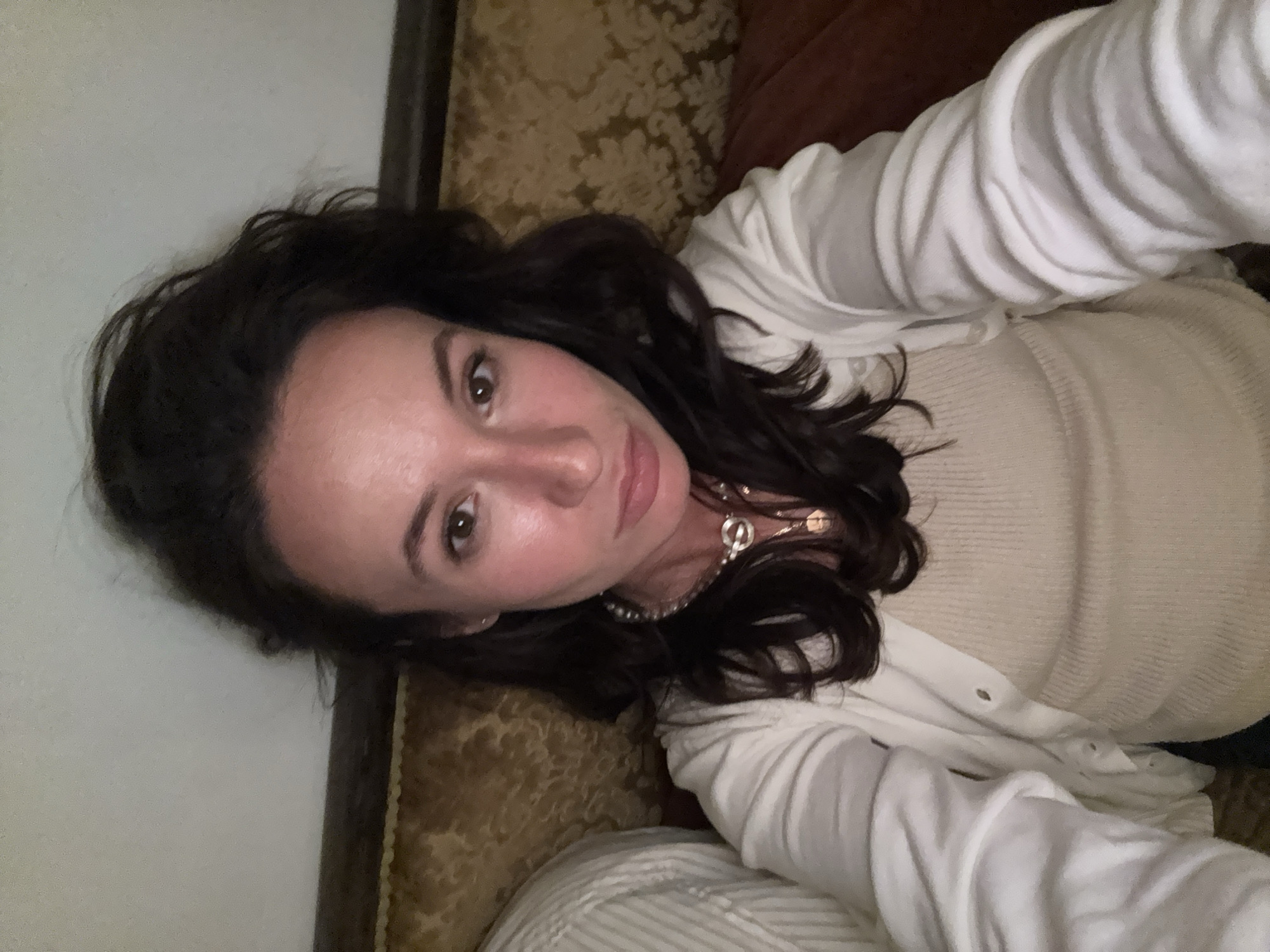 A woman with dark hair and fair skin is lying on a patterned couch. She is wearing a light-colored sweater and a white cardigan. She is looking at the camera. She is wearing a pearl necklace. The environment appears to be indoors. There is no information in the image to suggest that the location is Tel-Aviv, Israel.