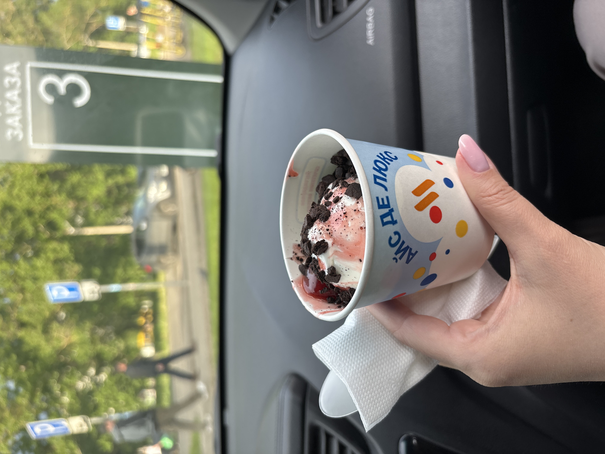 A person holds a cup of ice cream while sitting in a car in Rusanovo, Russia. The cup is white with blue and red designs and contains pink ice cream topped with chocolate cookie crumbles and a red sauce. A white napkin is under the cup. A sign displaying the number "3" with the text "ЗАКАЗА" partially visible is seen in the background through the car window. Trees and other blurred urban elements can also be observed outside the car. The car's dashboard and air vent are visible.