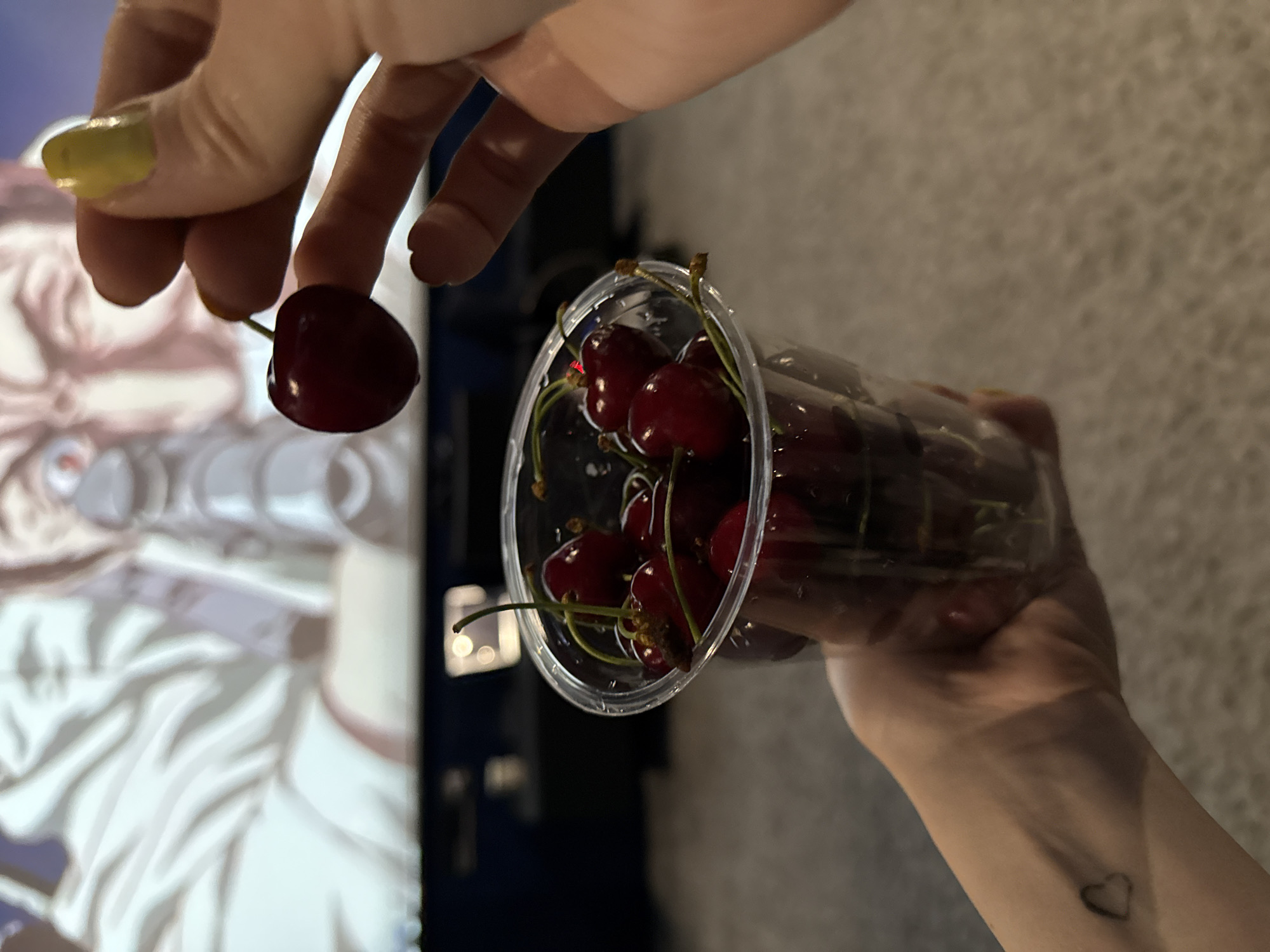 The image shows a person holding a clear plastic container filled with cherries. The person is also holding a single cherry between their fingers. The background includes a television screen displaying an image of a person and a textured wall. A small heart tattoo is visible on the person's wrist. The location cannot be determined from the image.