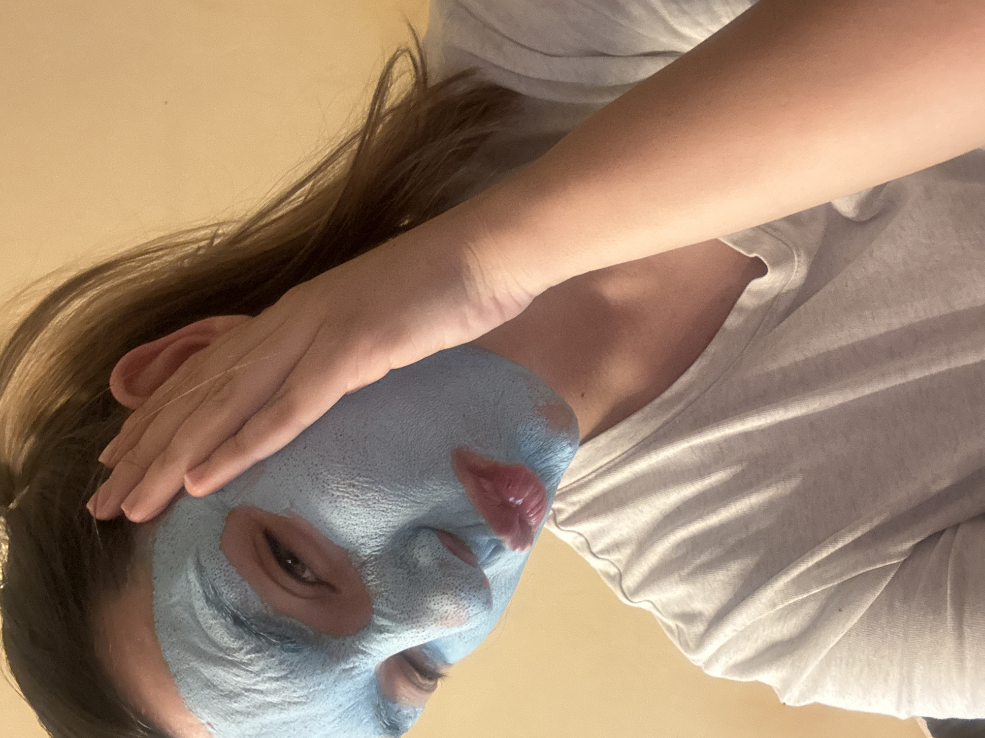 A person with a light blue face mask on their face is positioned in the center of the image. The mask covers the entirety of their face, with openings left for the eyes, nose, and mouth. The mask appears to be cracked. The person is wearing a white T-shirt. Their hand is resting on the upper portion of their head. The background is a light cream color. There is no evidence to suggest this image was taken in Sofia, Bulgaria.