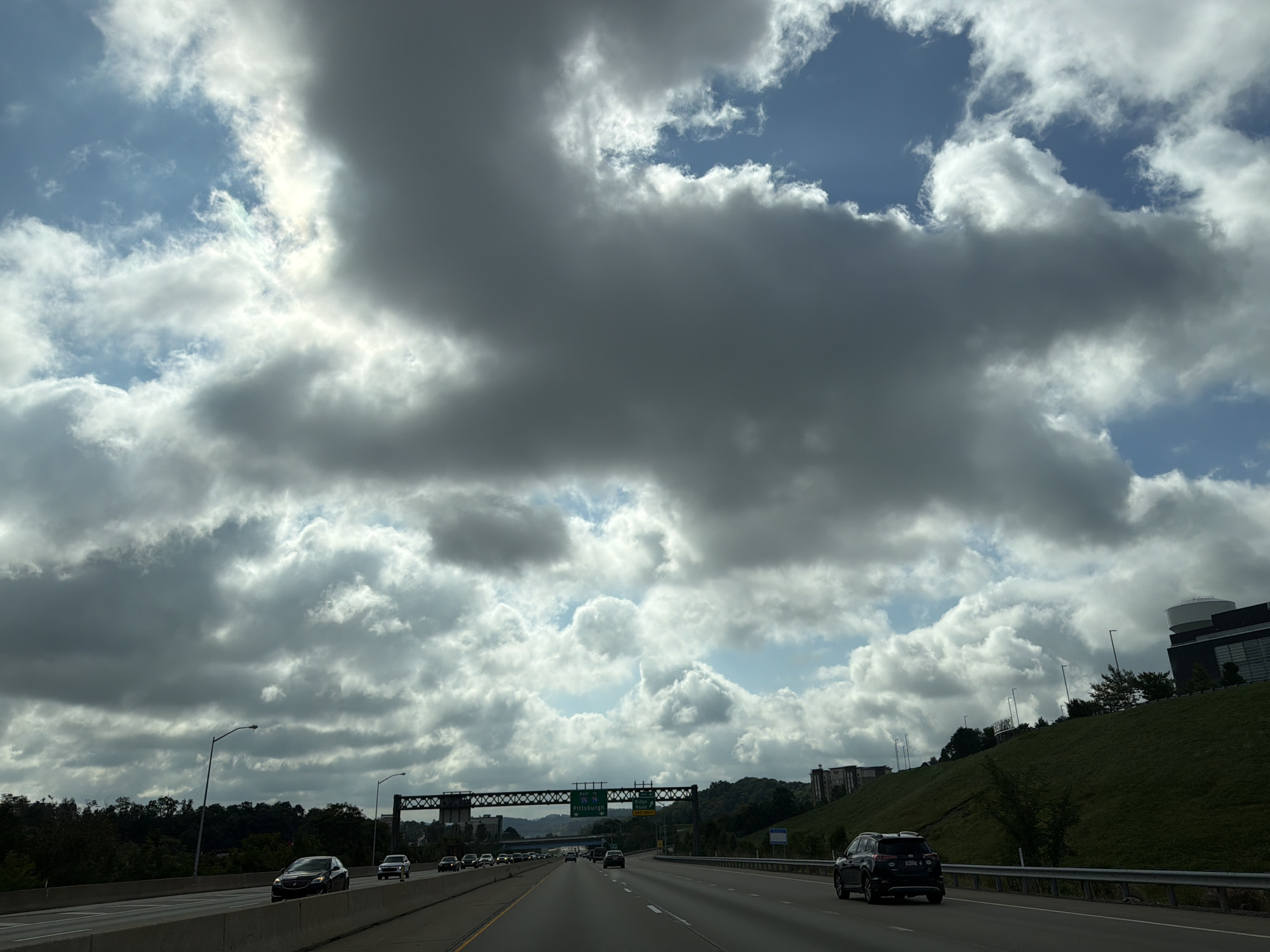 This is a wide shot captured from a vehicle driving on a highway in Robinson Township, United States of America. The sky is filled with dramatic, heavy clouds, with patches of blue sky peeking through. The clouds are a mix of dark gray and bright white, indicating a dynamic weather system.

The highway is multi-lane and bordered by concrete barriers. Several vehicles are visible on the road, including a black SUV in the foreground to the right, and a black sedan in the middle lane of the highway. On the left side of the highway, there is a grassy embankment with trees and streetlights. In the distance, a large overpass with signs is visible. Further down the highway, more cars are driving. On the right side of the highway, beyond the grassy hill, there are several modern buildings. The overall scene suggests a drive on a cloudy day.