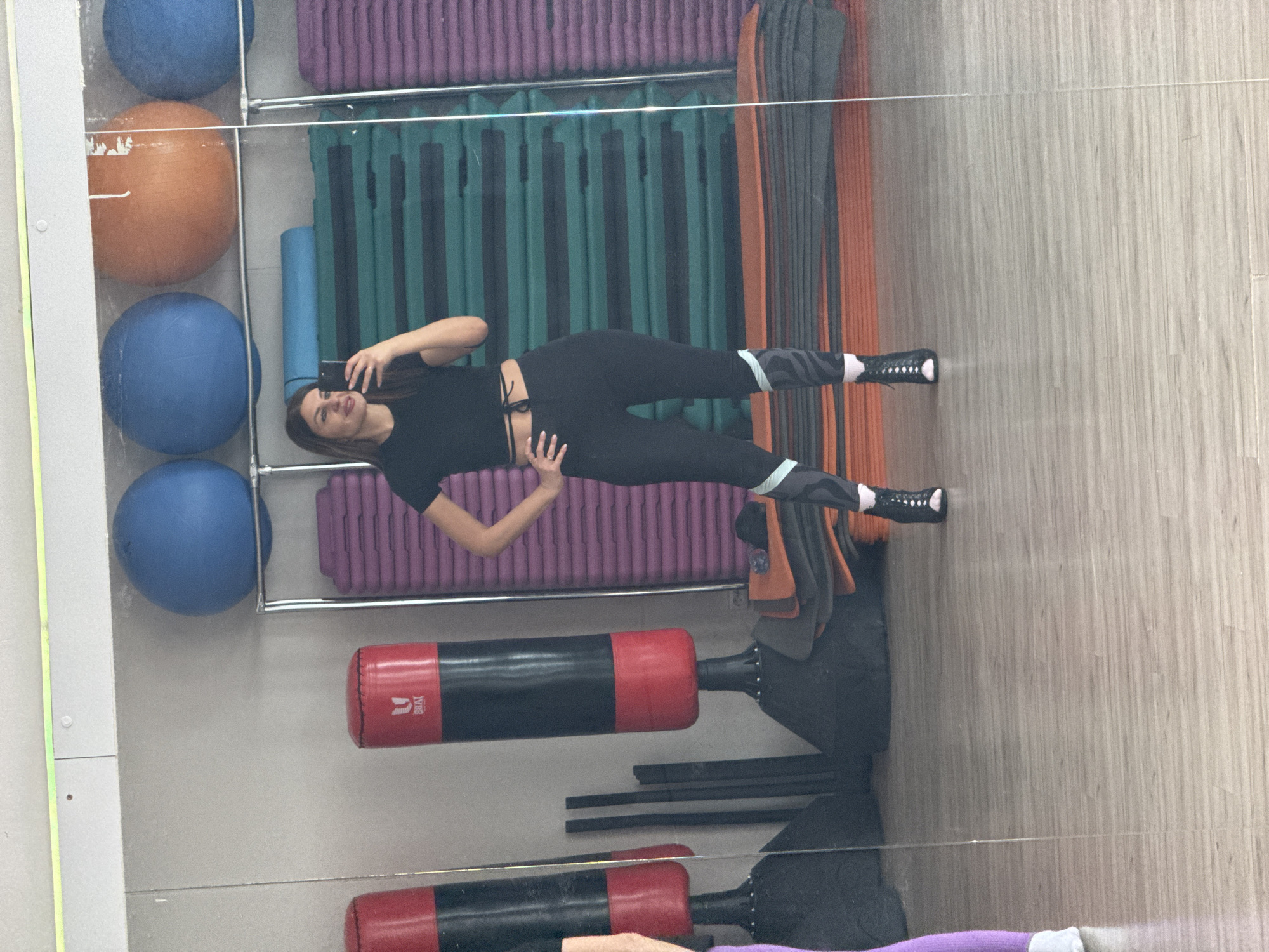 The image appears to be a reflection in a mirror in a fitness center. A woman in black athletic clothing is holding a smartphone, presumably taking a selfie. Various fitness equipment is visible, including exercise balls, step platforms, punching bags, and mats. The location is likely Chaikovsky, Russia, given the query context.