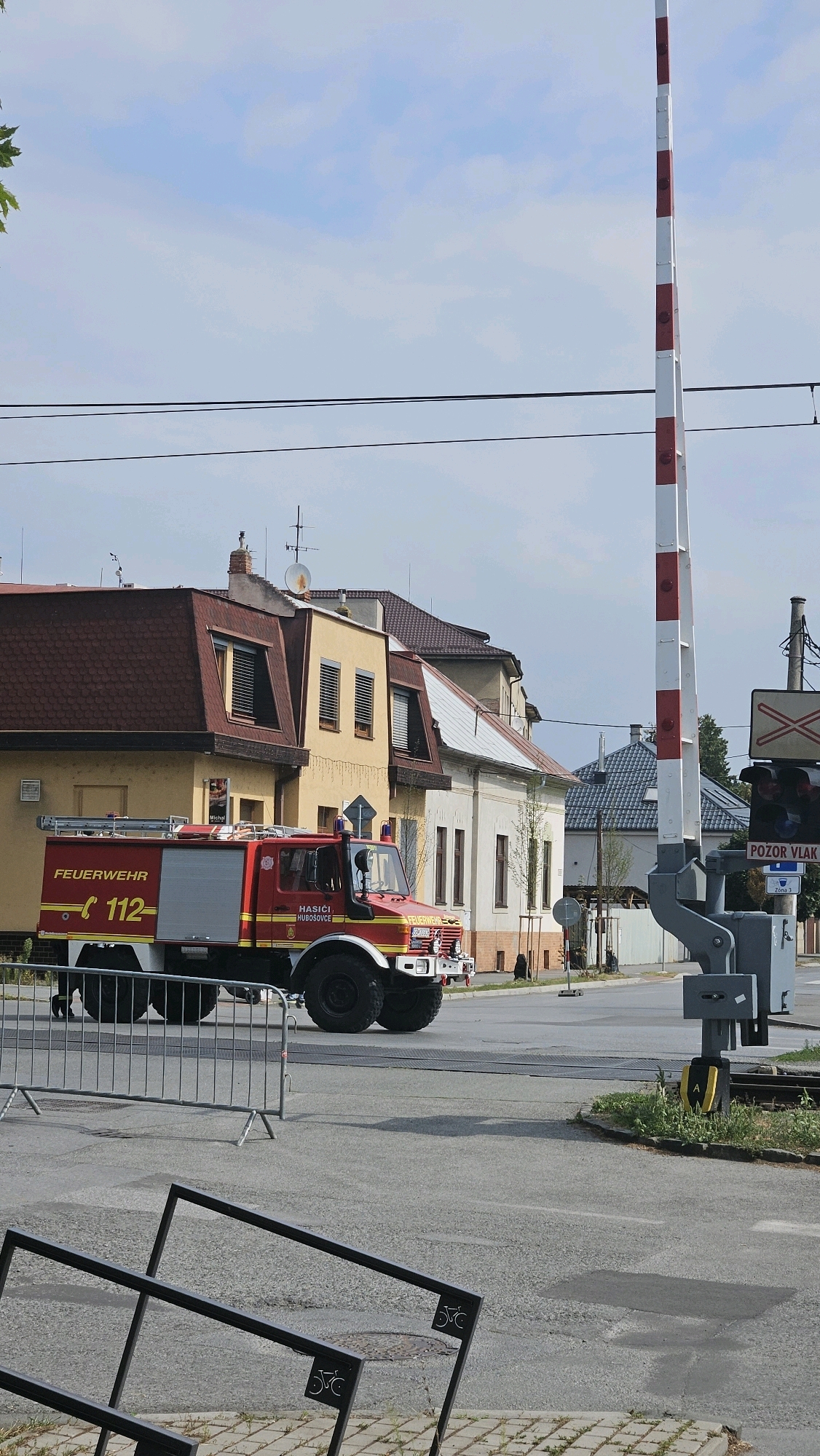 A red fire truck with "FEUERWEHR" and "112" written on its side is parked on a street. The fire truck has "HASICI HUBOŠOVCE" written on the side of the cab. The fire truck is facing left, with its front wheels slightly turned. Behind the fire truck are several buildings with tiled roofs. A railway crossing signal with red and white stripes is to the right of the fire truck. The sky is cloudy. The image appears to be taken on a street in the city of Prešov, Slovakia, likely near a residential area due to the presence of the houses.
