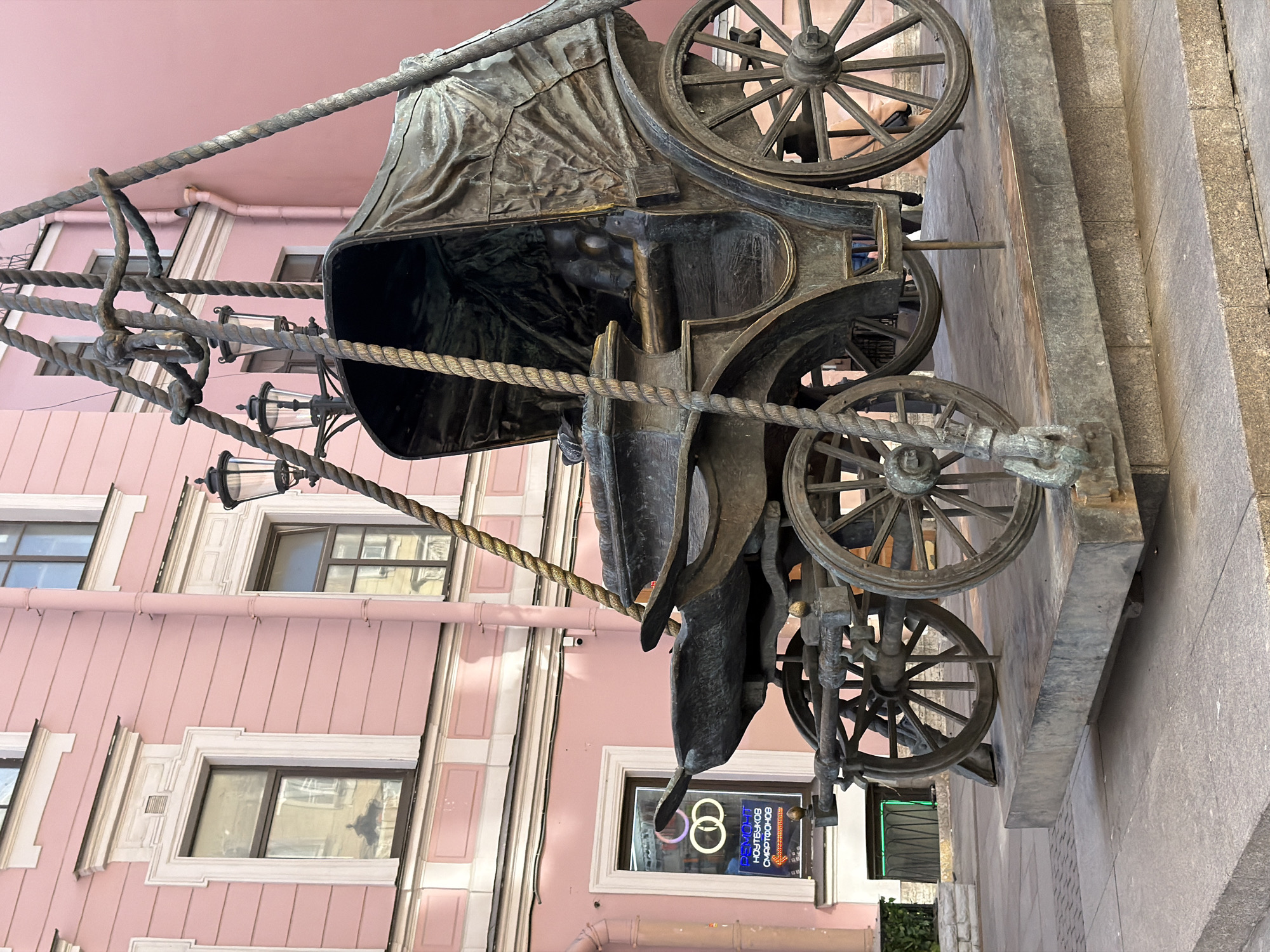 The image shows a bronze statue of a horse-drawn carriage with four wheels. Two lamps hang from ropes connected to the carriage. The statue is placed outside on a sidewalk next to a pink building with multiple windows. A storefront is visible through a window on the bottom floor of the pink building. The image is from the city of Saint Petersburg, Russia.