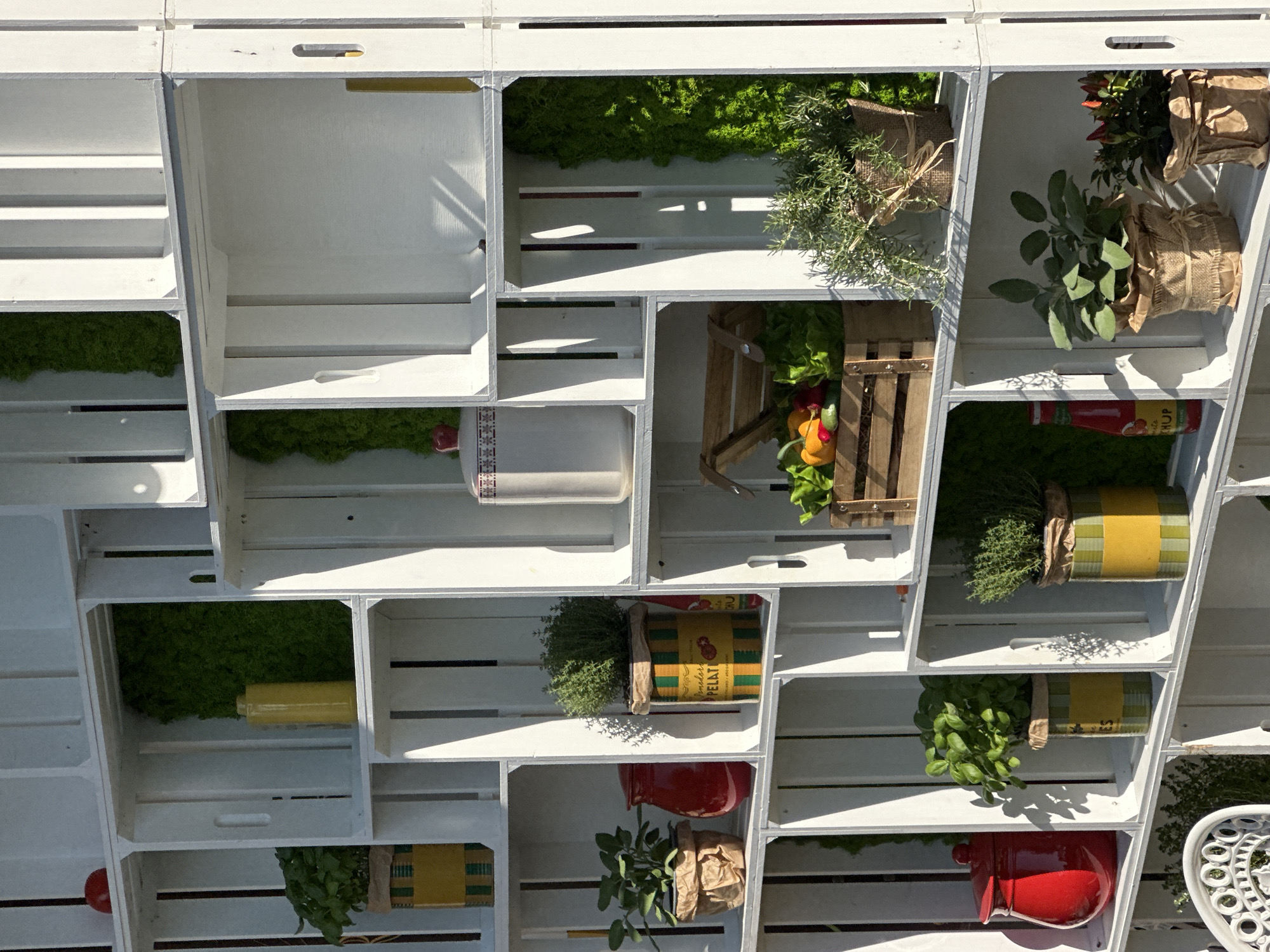 The image displays a white shelving unit constructed from wooden crates, filled with various plants and decorative items. The shelving unit is set against an outdoor backdrop, suggesting it is part of a garden or an outdoor market stall. The crates are arranged in a grid pattern, creating individual compartments.
In the top row, on the left, a red object, possibly an apple, rests on top of a crate. The second crate from the left contains green moss and a yellow ceramic vase. The third and fourth crates are empty.
The second row from the top features a plant in a yellow and green pot on the left, a red ceramic jar next to it, and another plant in a similar yellow and green pot. To the right, a white ceramic container with a red lid and decorative patterns is visible.
The third row contains a wooden crate filled with fresh vegetables, including lettuce, yellow bell peppers, and red bell peppers. To the right of this crate is a potted herb plant with green leaves, wrapped in burlap and tied with twine. Further to the right, there's another compartment with green moss.
In the bottom row, on the left, a red jar is placed next to a potted herb plant in a yellow and green container. To its right, another potted herb plant in a yellow and green container is visible. Further to the right, a bottle of ketchup is standing upright. On the far right, a potted plant with small red peppers is present.
The lighting suggests it is daytime, with sunlight casting shadows on the shelving unit and its contents. The overall impression is one of a rustic and decorative display, likely intended for a garden center, a farmer's market, or an outdoor cafe. The mention of Varenna, Italy, as the location implies this scene is situated in a picturesque Italian town, possibly along the shores of Lake Como, known for its charming streets and vibrant floral displays. However, there are no specific visual cues within the image that directly confirm this location.