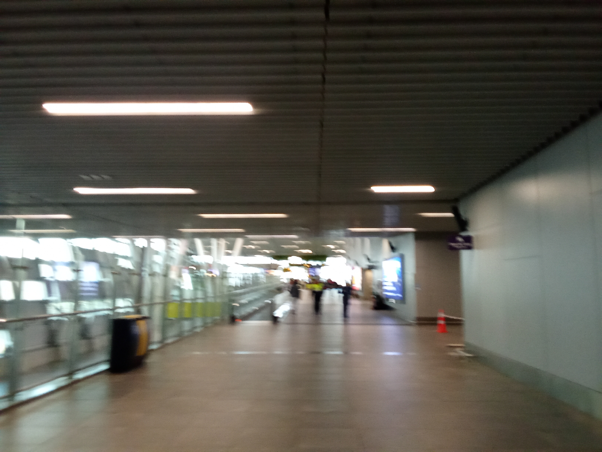 The image depicts an indoor walkway, possibly within an airport terminal or a large public building, characterized by a modern, industrial design. The ceiling is a corrugated metal structure with recessed rectangular lighting fixtures providing bright, uniform illumination. The floor is a polished, light-colored material, reflecting the overhead lights.

On the left side of the walkway, a transparent glass barrier runs along the length of the corridor, revealing a glimpse of an escalator or moving walkway behind it. A dark cylindrical trash receptacle with a yellow band is positioned next to the glass barrier. Further down the walkway, several blurred figures are visible, suggesting people are walking in both directions. Some individuals appear to be wearing reflective vests, hinting at a possible work or security function.

On the right side, a plain, light-colored wall extends along the corridor. Mounted on this wall is a small, dark sign with a purple logo. Further down the right wall, a brightly colored orange traffic cone is placed on the floor, indicating a potential obstruction or a demarcated area. A large, illuminated digital display or advertisement is visible on the far right wall. The overall impression is one of a functional, contemporary public space, with a focus on efficient movement and clear lighting. The specific location is Pudahuel, Chile, which suggests this is likely within the Arturo Merino Benítez International Airport.