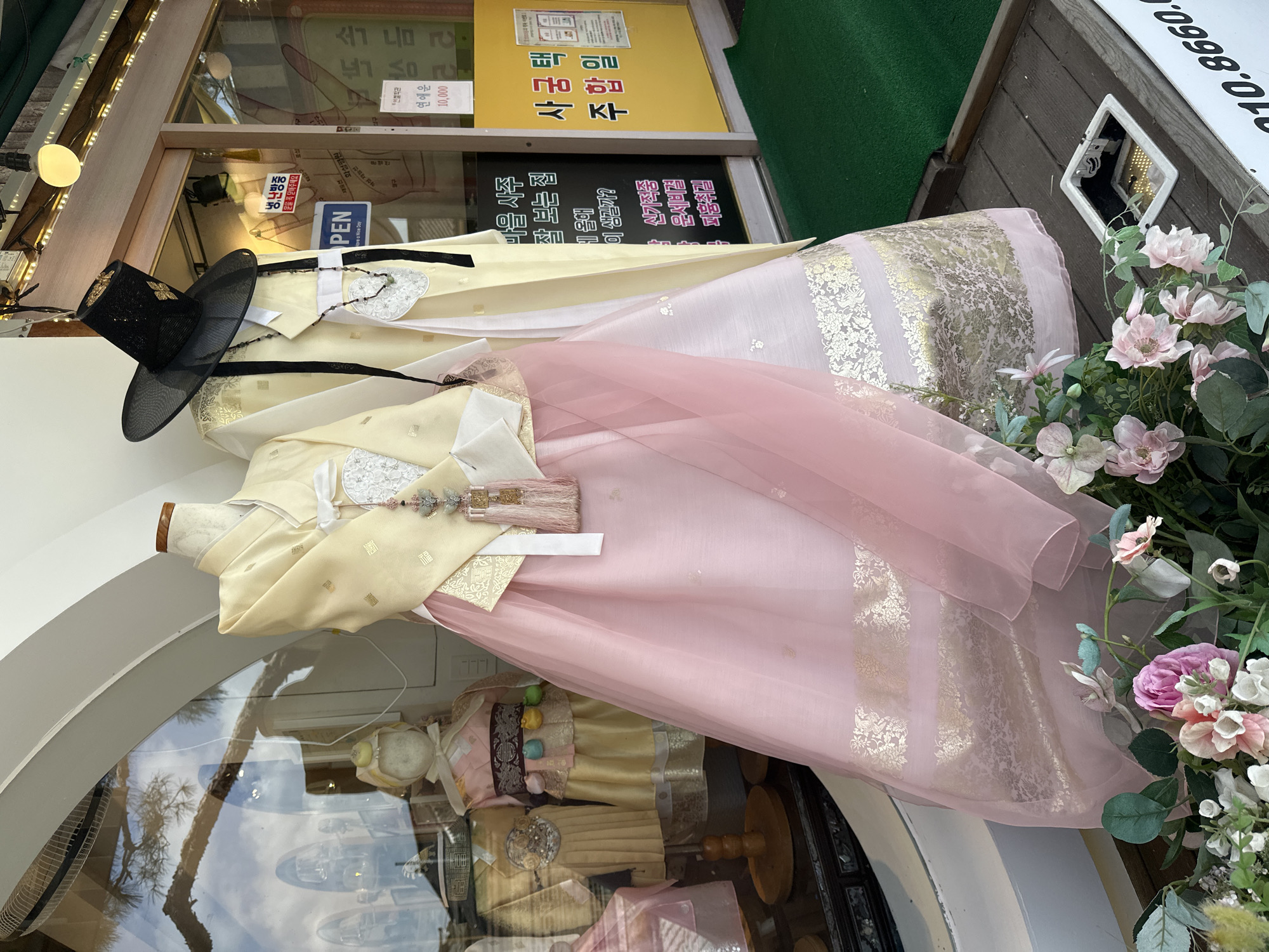 This image depicts a storefront display featuring traditional Korean clothing, likely Hanbok, in Jeonju-si, South Korea. The display is set up outside, possibly on a wooden deck, with a reflective window in the background. Two mannequins are prominently displayed, one wearing a pale yellow jeogori (top garment) with intricate gold patterns and a flowing pink skirt, and another mannequin wearing a similar yellow jeogori with a black traditional hat. The garments are adorned with decorative tassels and jewelry. In the background, through the window, other traditional clothing items and mannequins can be seen. There are also various signs in Korean text on the storefront. Several potted artificial flowers are placed at the bottom of the display, adding a touch of color and softness. A white sign with a phone number is visible along the bottom edge of the image. The overall scene suggests a shop selling or renting traditional Korean attire in a tourist-frequented area of Jeonju, known for its well-preserved traditional Korean villages.