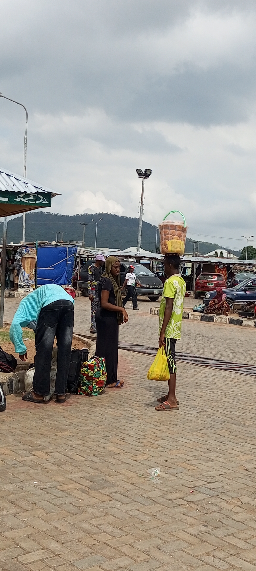The scene is located in Lokoja, Nigeria, depicting an outdoor area paved with interlocking bricks. To the left, a person in a light blue long-sleeved shirt bends over luggage. A woman in a black dress and olive-green headscarf stands near a patterned tote bag. A man stands directly across from her, wearing a lime-green shirt with black shorts, holding a yellow plastic bag and carrying a bucket of bread on his head. In the background, there are several parked cars, street vendors under makeshift shelters, and people moving around. A mountain is visible in the distance beneath a cloudy sky. A tall light pole is also present in the scene.