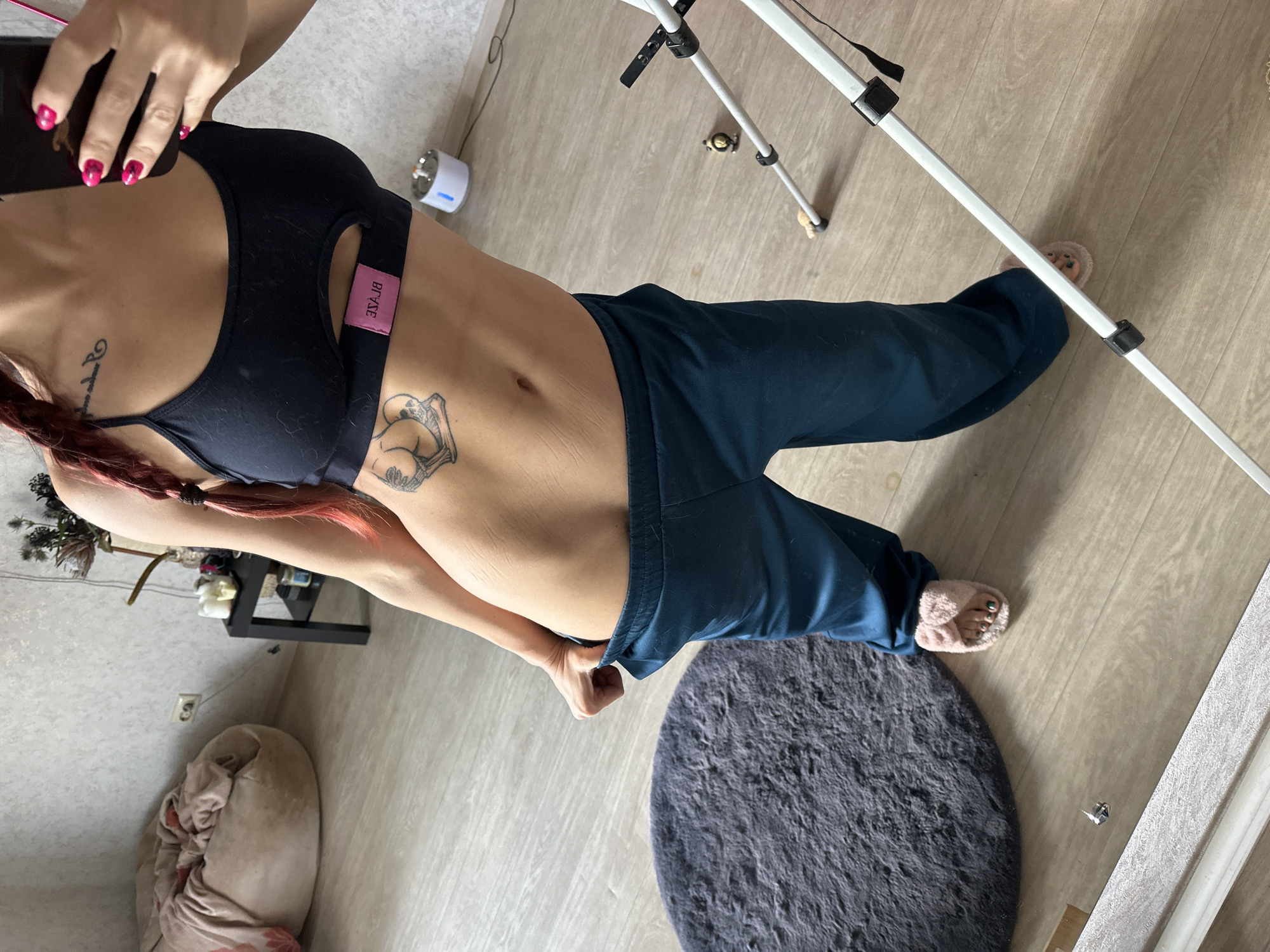 A woman wearing a black sports bra and blue pants stands in a room. The room has light wood-patterned flooring, a grey circular rug, and various objects including a tripod, a black shelf unit, a beanbag chair, and a white appliance. The woman has a tattoo on her abdomen and another on her chest. The location is not specified, and there is no evidence it is in Vladimir, Russia.